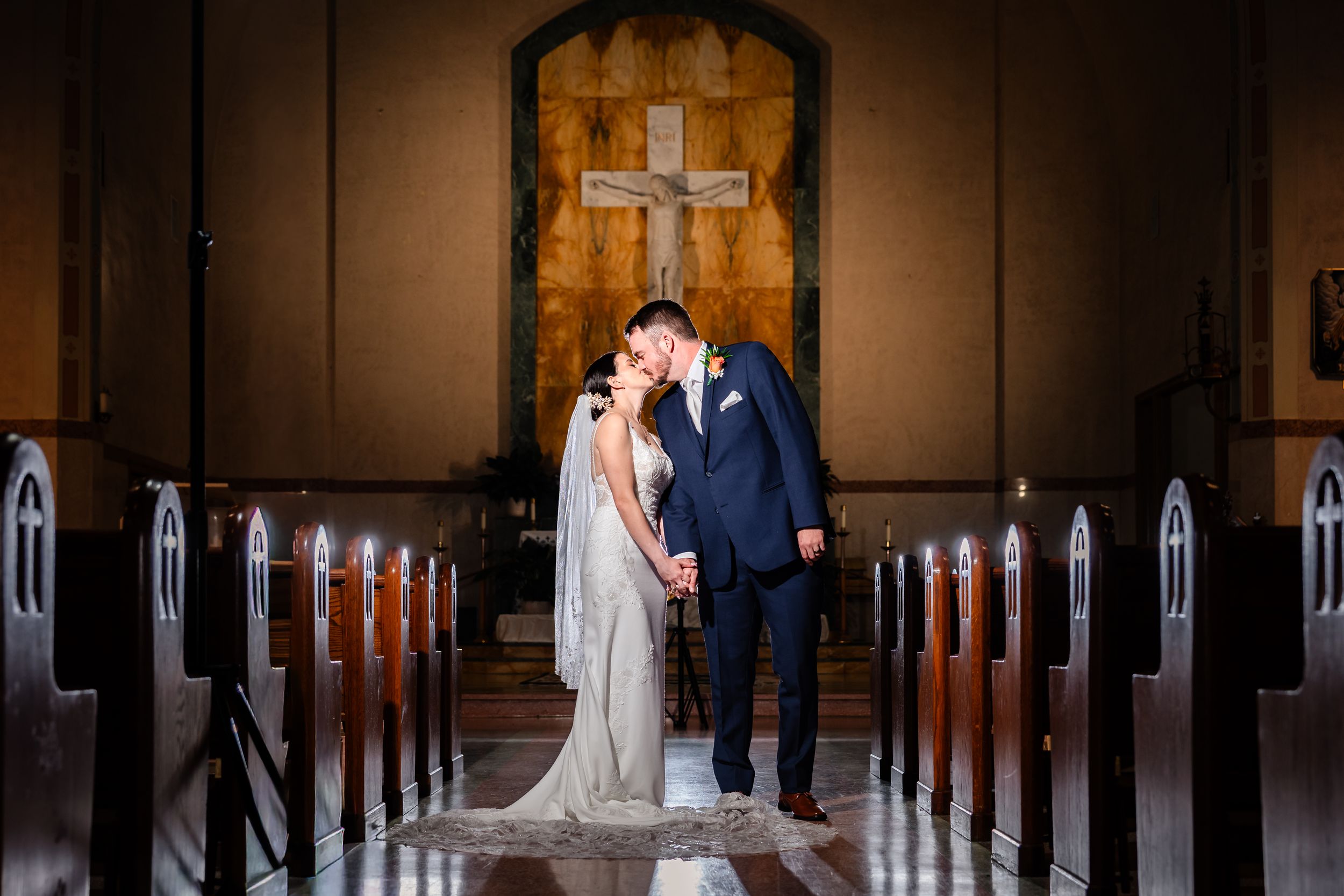Bride and groom sharing a romantic kiss in a church aisle with a cross backdrop.