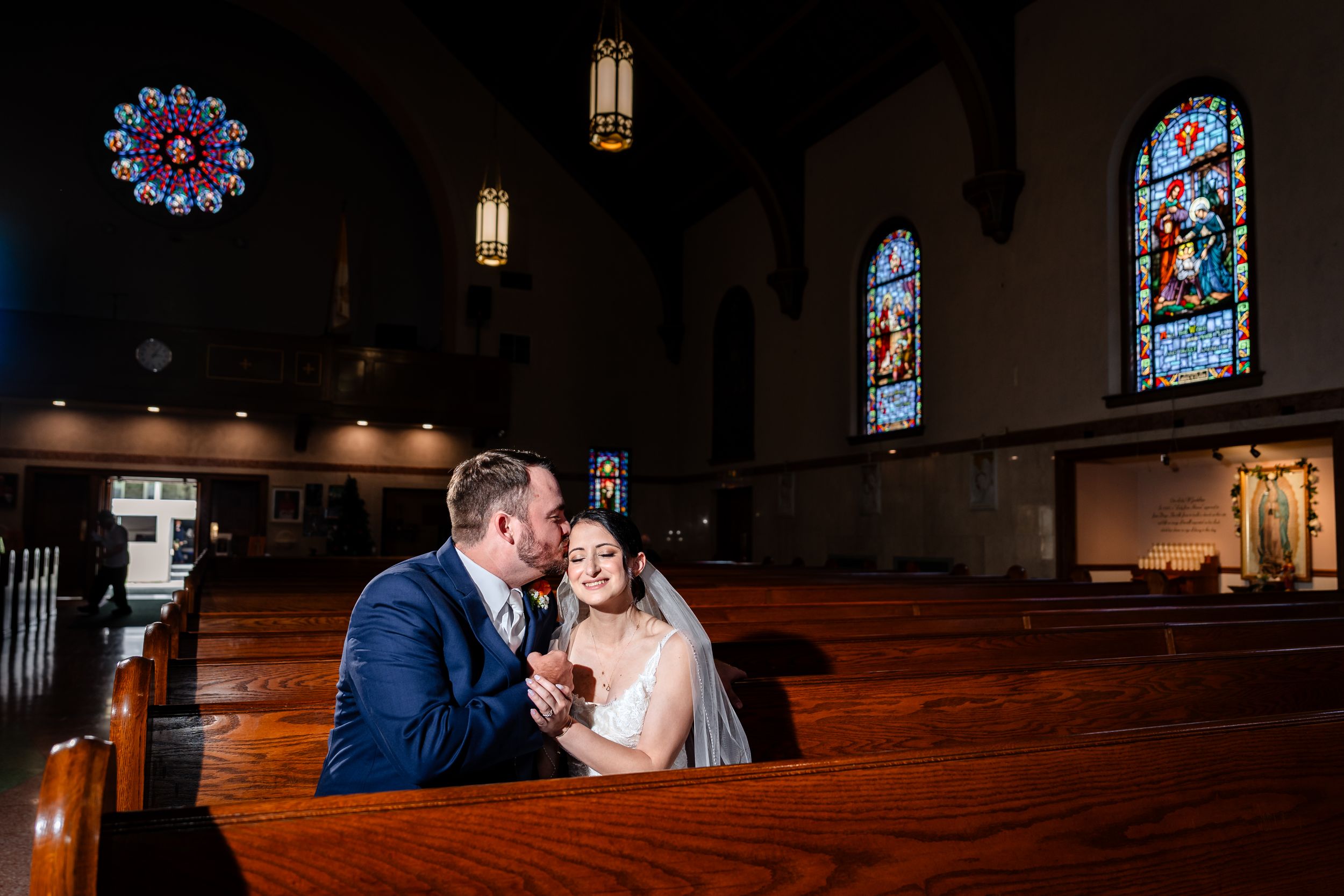Bride and groom share a tender moment in a church pew surrounded by stained glass windows on their wedding day.