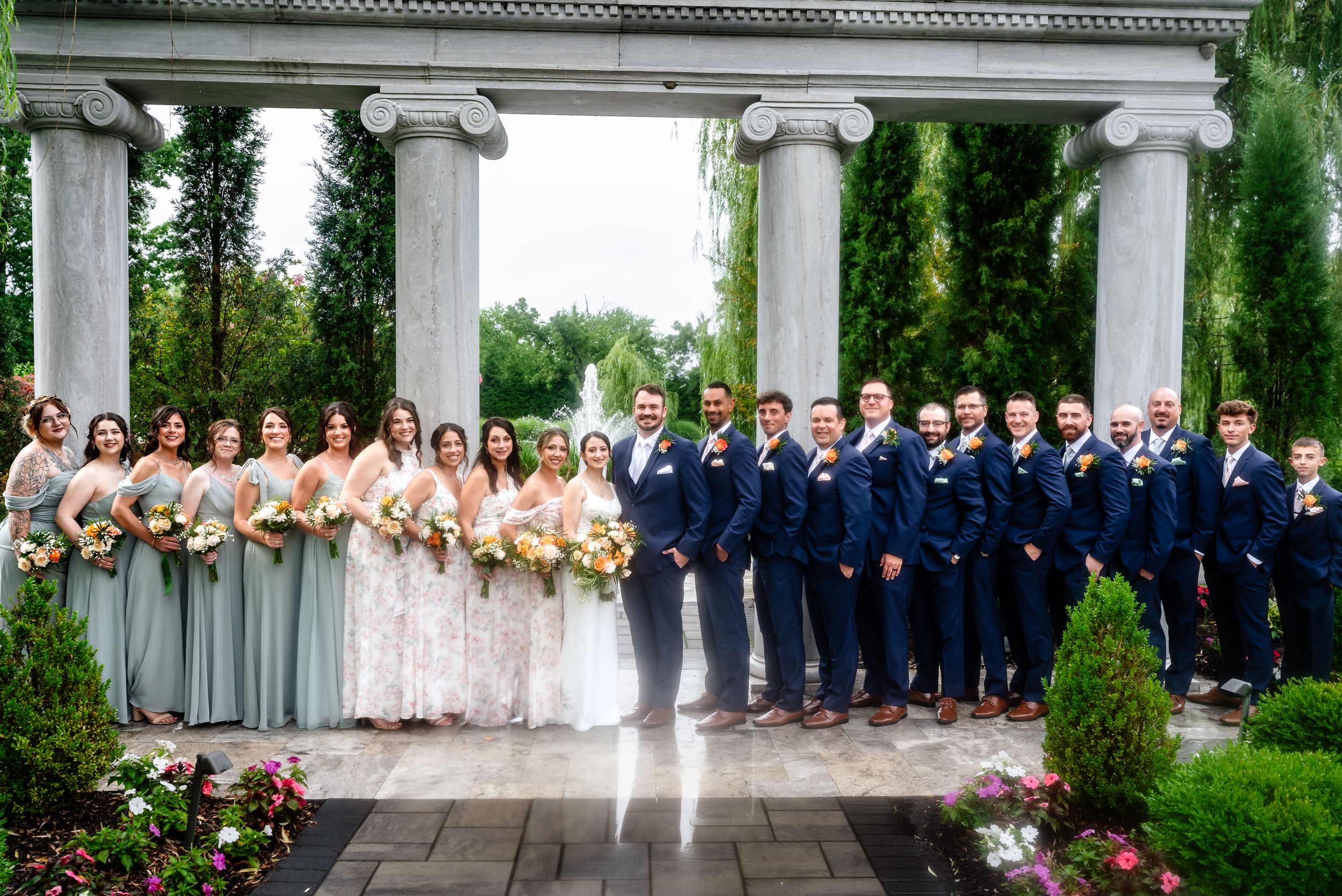Wedding party posing under a stone pergola with fountains and lush greenery outdoors.