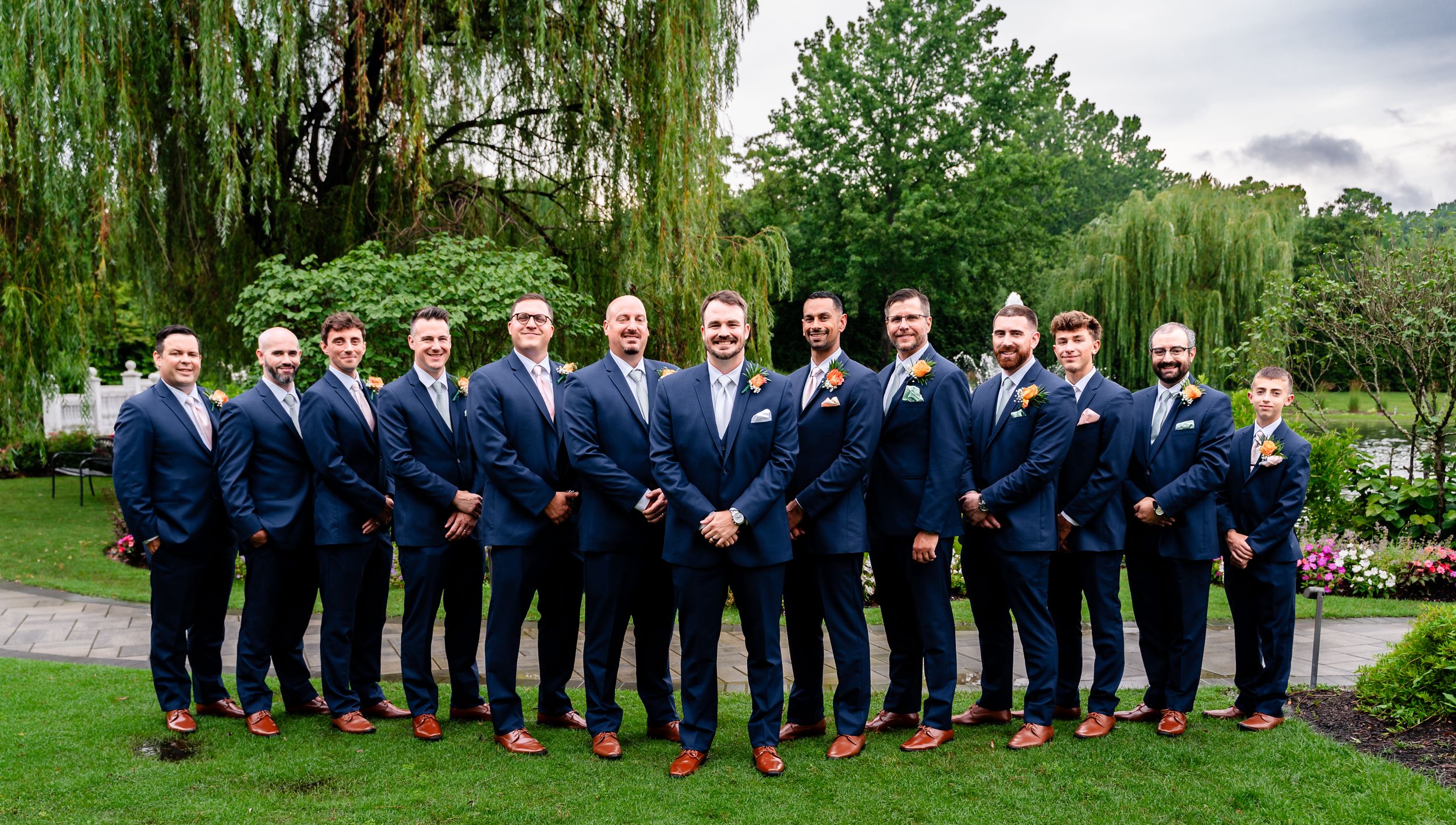 Groom and groomsmen pose in matching navy suits with red shoes in a lush garden setting.