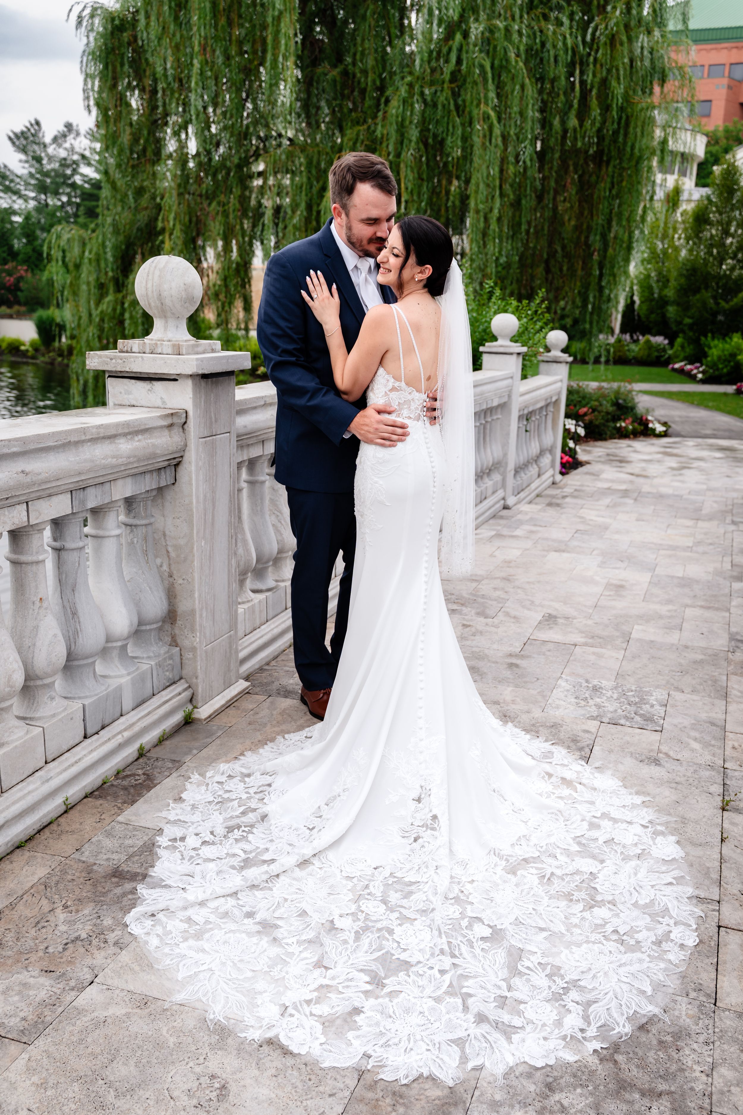Bride and groom embracing on stone bridge outdoors, bride wearing elegant lace train wedding dress.