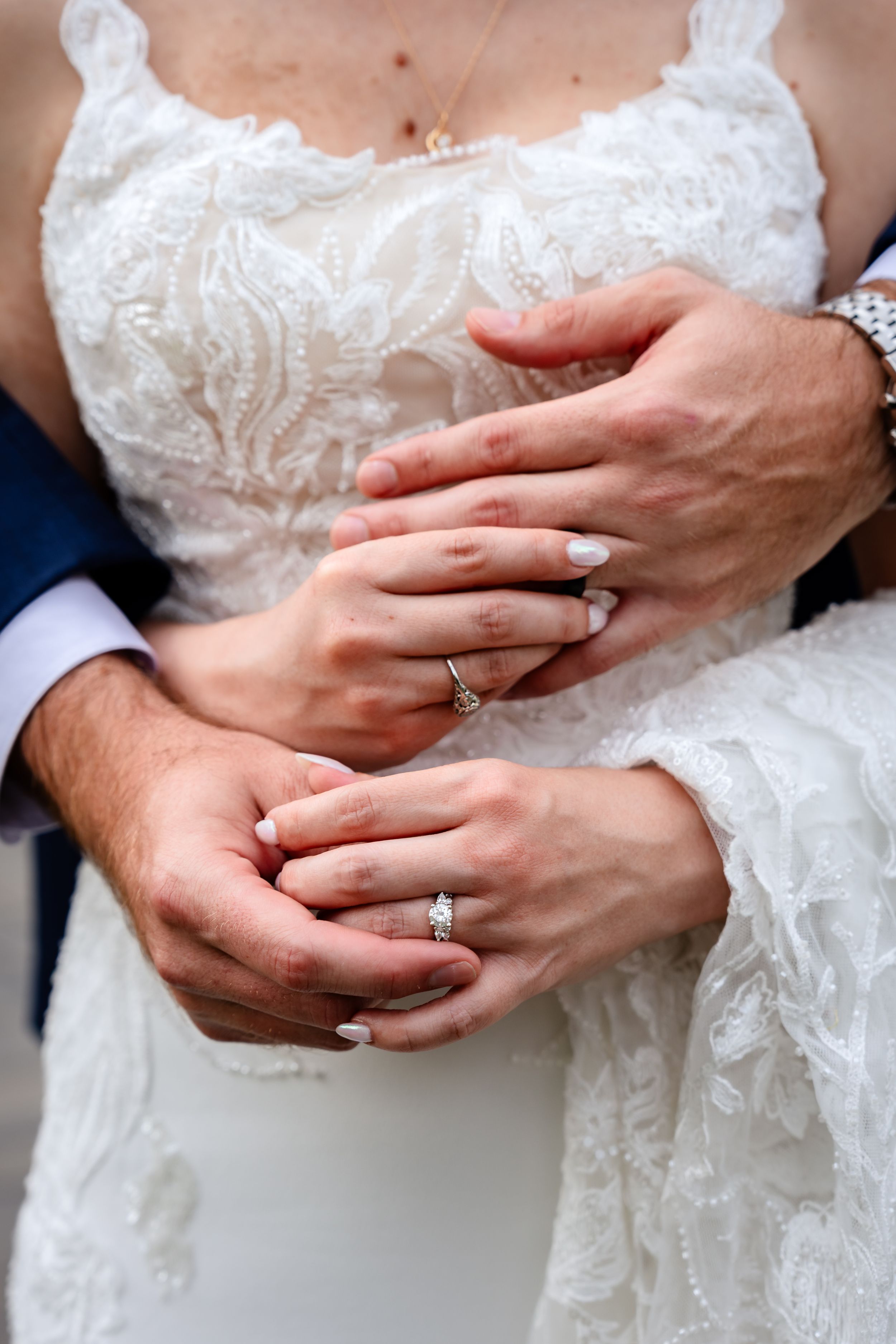 Close-up of bride and groom hands displaying wedding and engagement rings on white lace gown.
