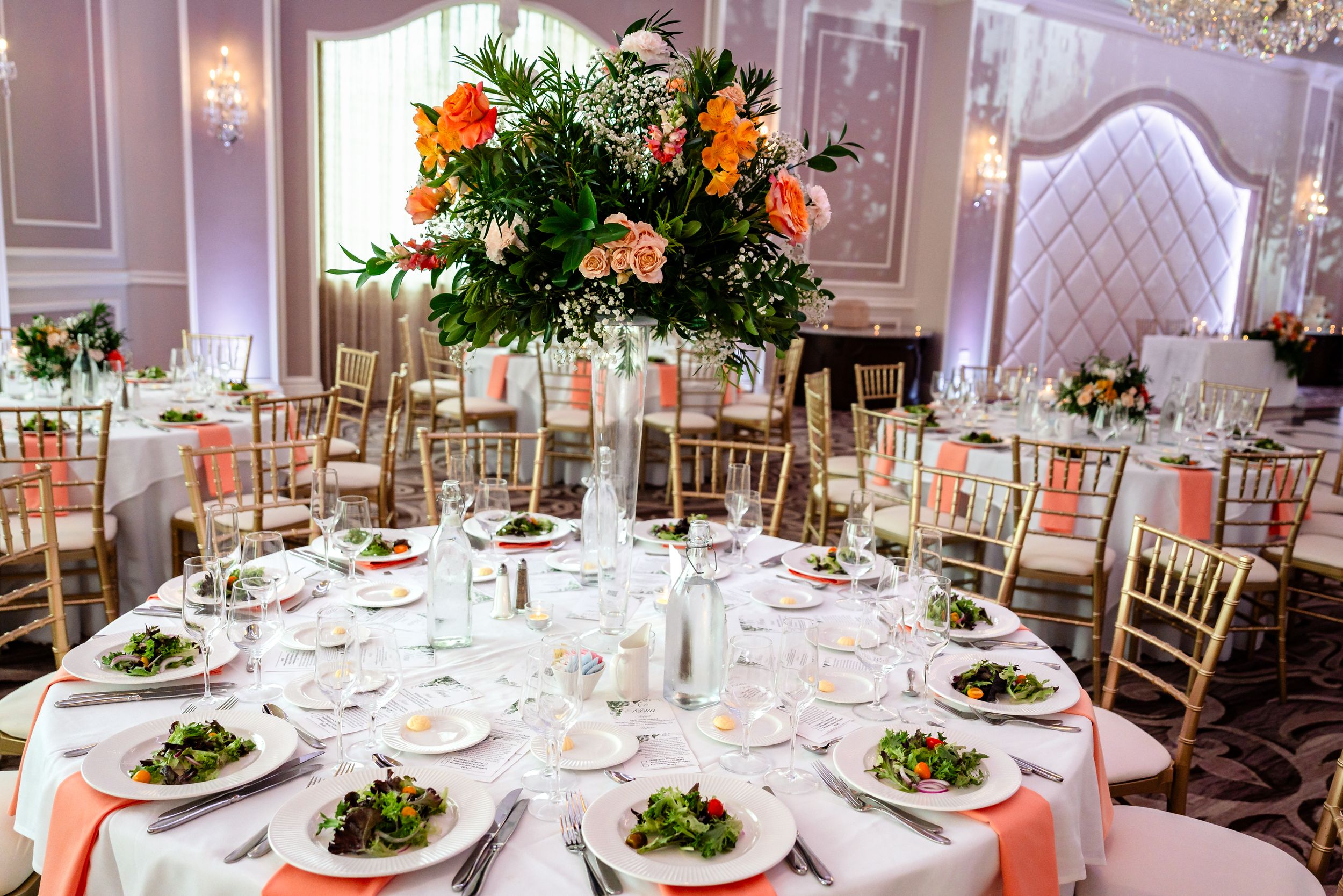 Elegant wedding reception table with peach linens, gold chiavari chairs, and a tall floral centerpiece of orange blooms and greenery.