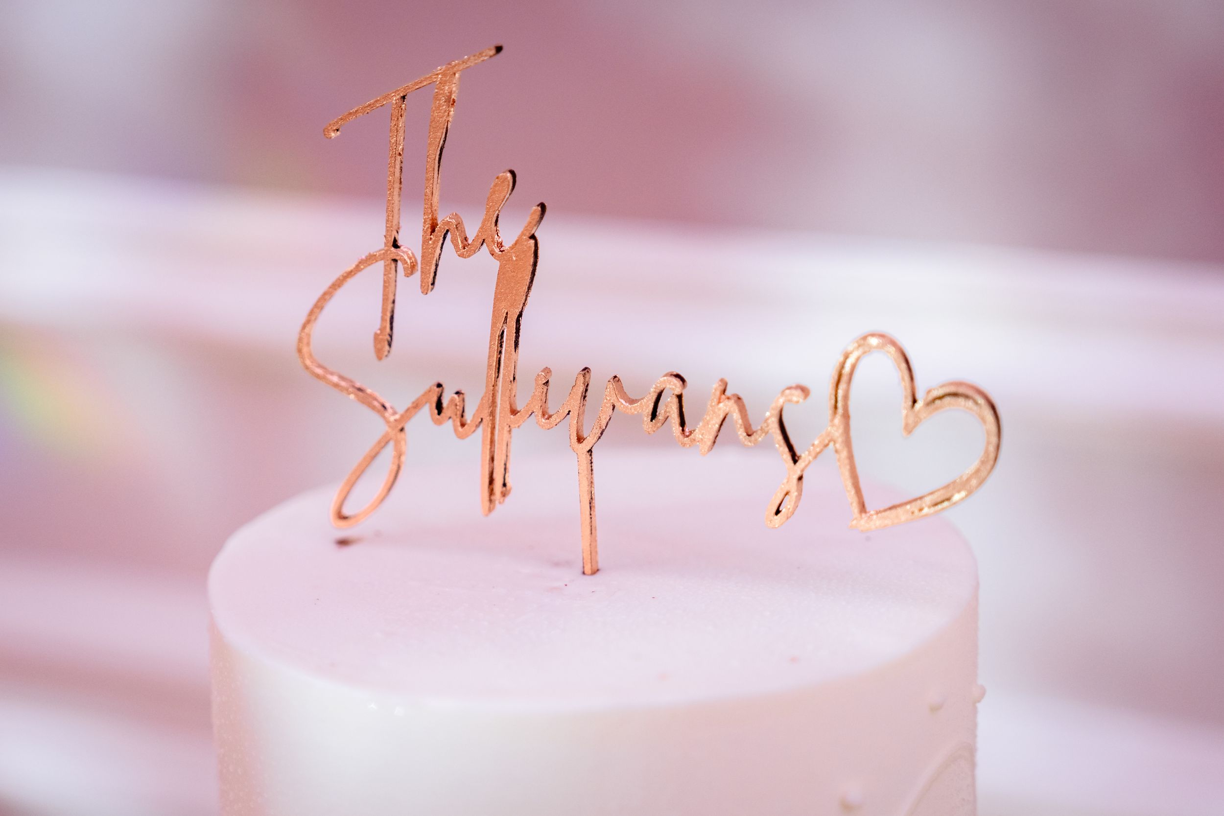 Rose gold acrylic cake topper reading The Sullivans with a heart detail on a white wedding cake.