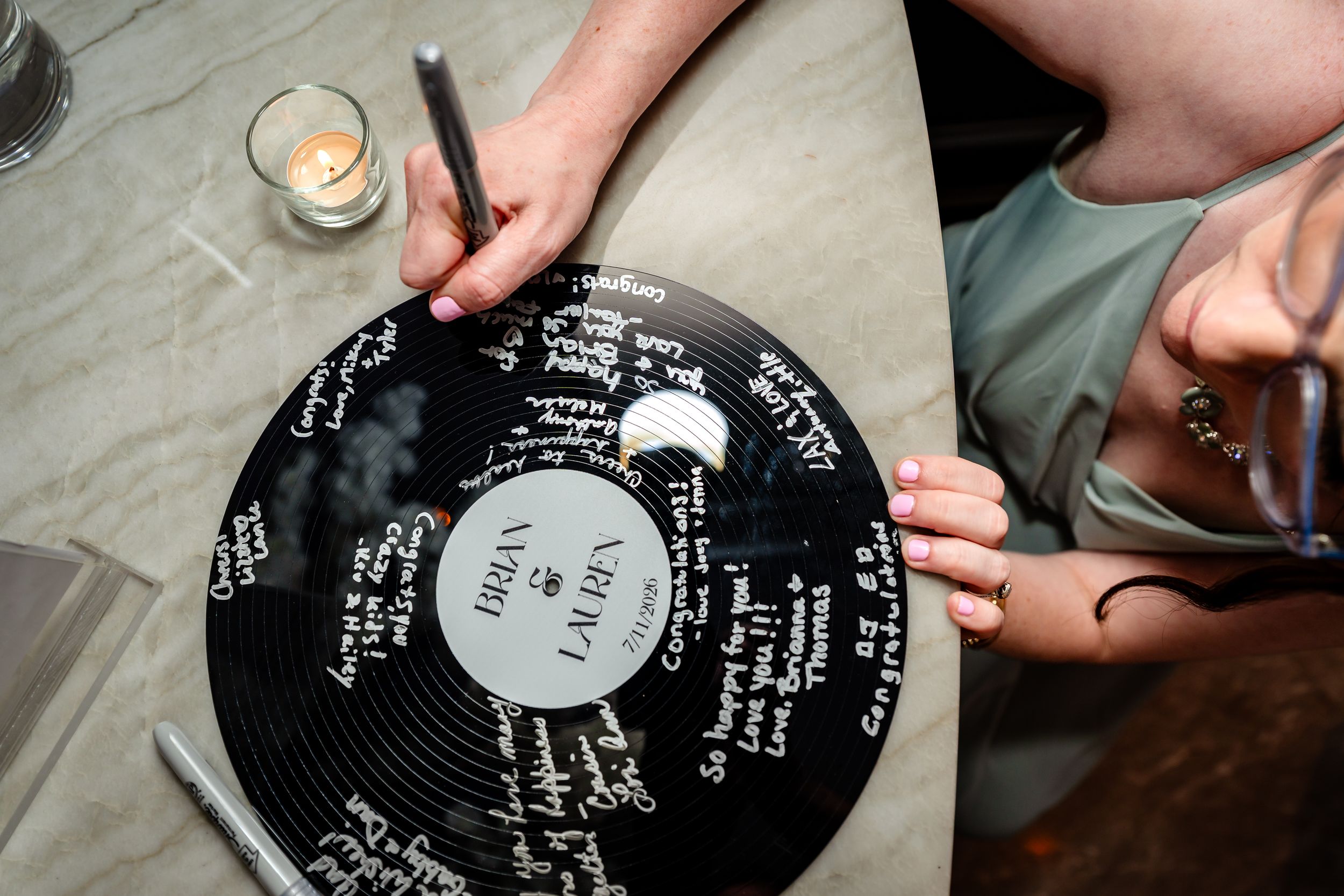 Guests signing a vinyl record wedding guestbook with white marker at a reception table with candles.