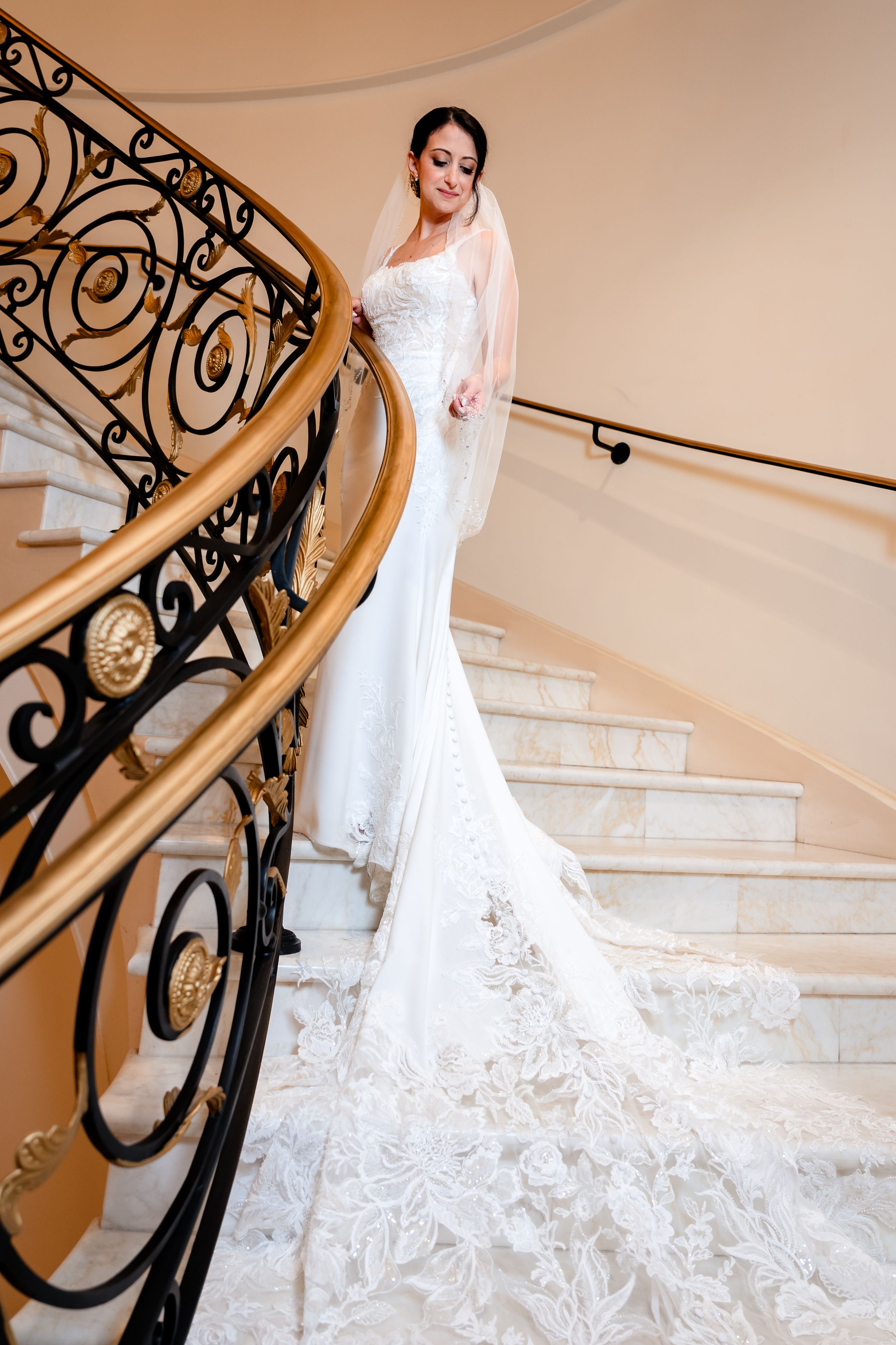 Elegant bride in fitted lace wedding gown with long train posing on ornate spiral staircase.