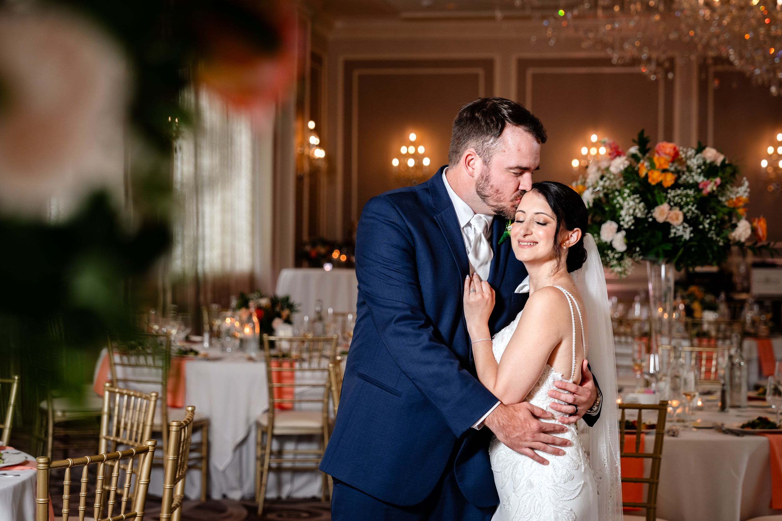 Groom embracing bride in elegant reception hall with chandelier lighting.