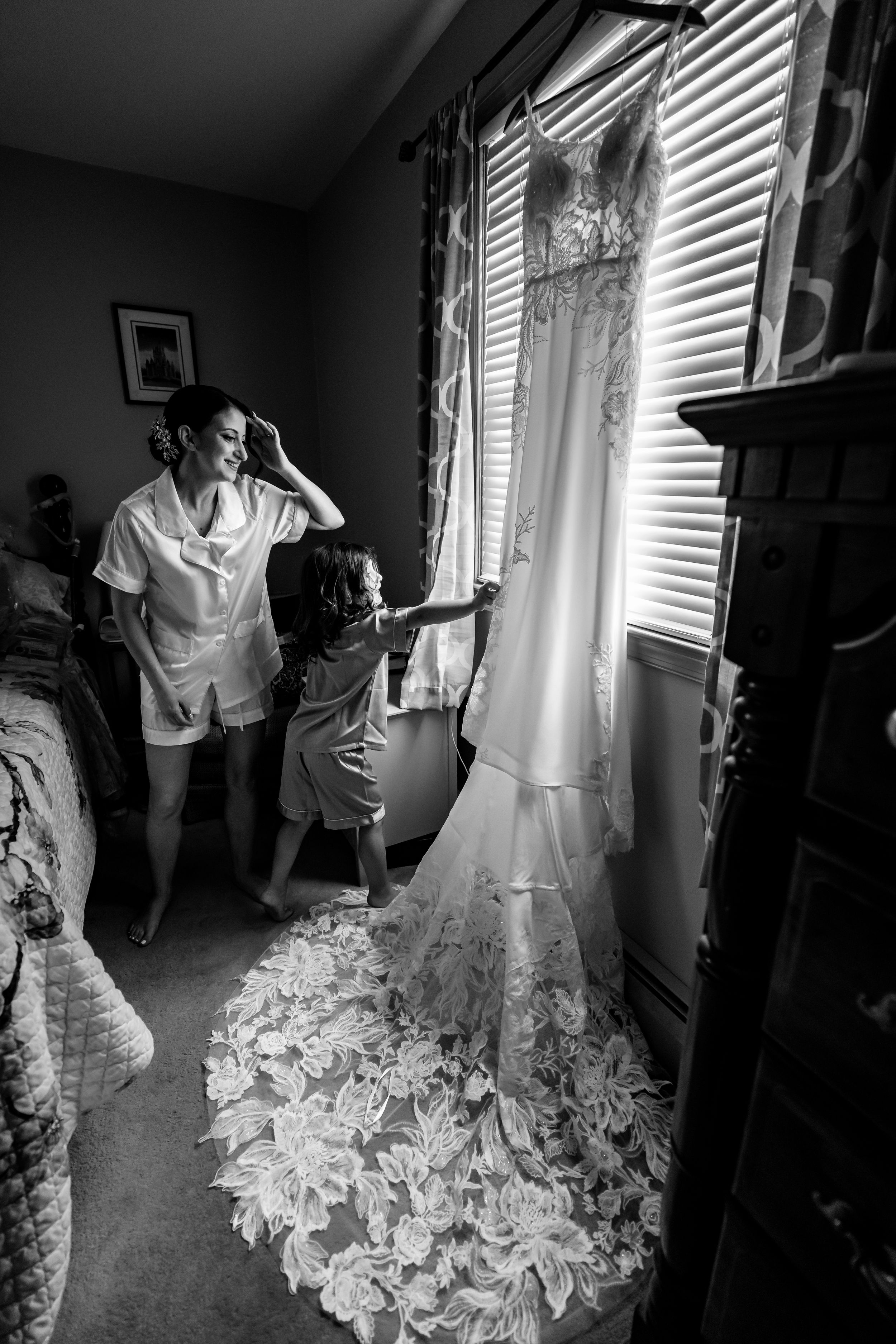 Black and white photo of bride admiring her lace wedding dress hanging by window with child.