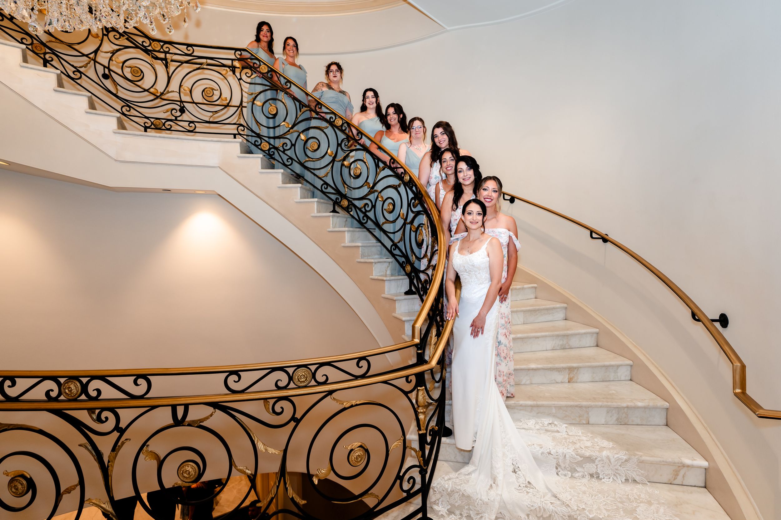 Bride in white gown poses with bridesmaids on elegant curved staircase with ornate iron railing at wedding venue.