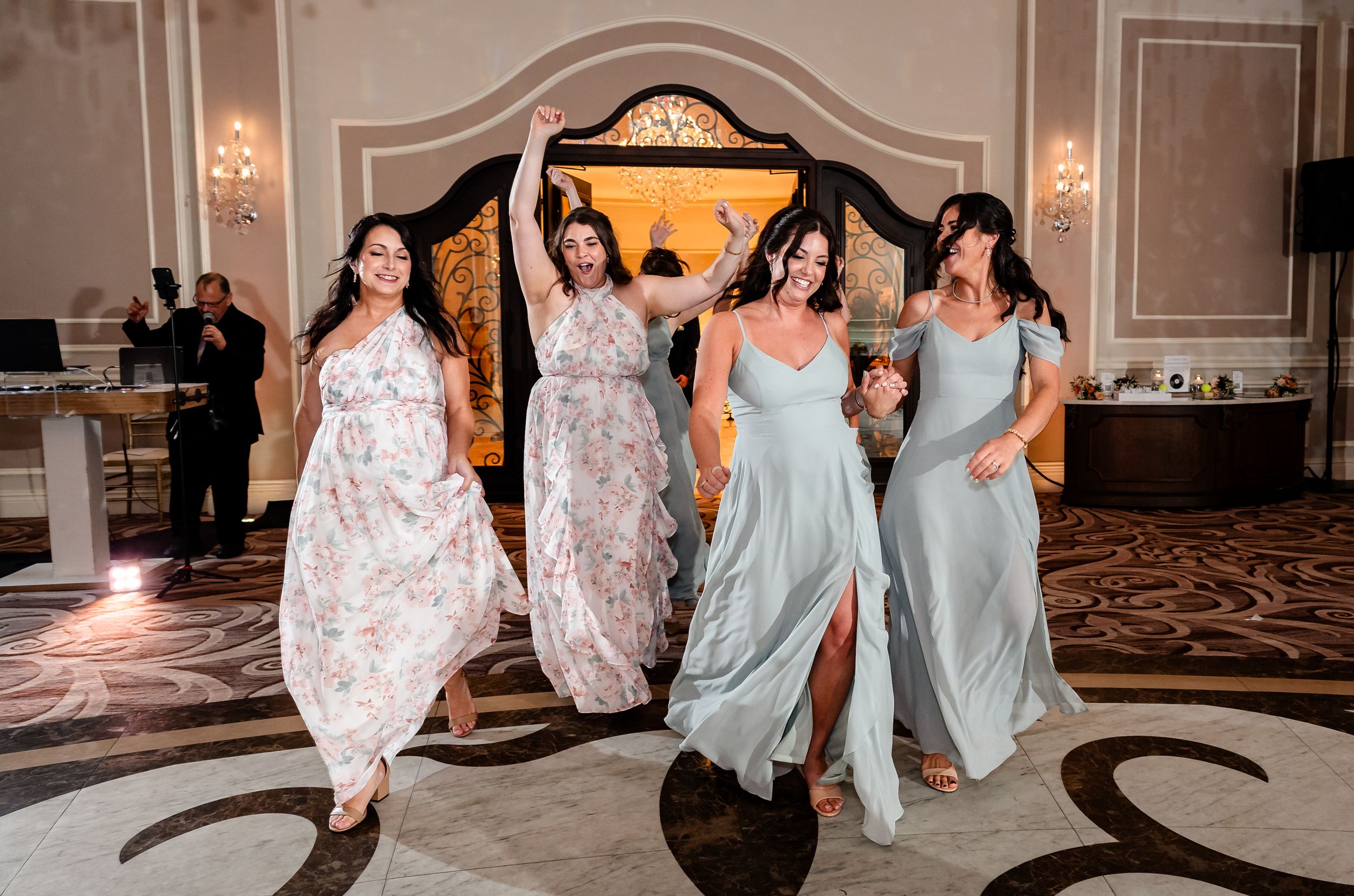 Four bridesmaids in elegant gowns dancing joyfully at a wedding reception in a grand ballroom.