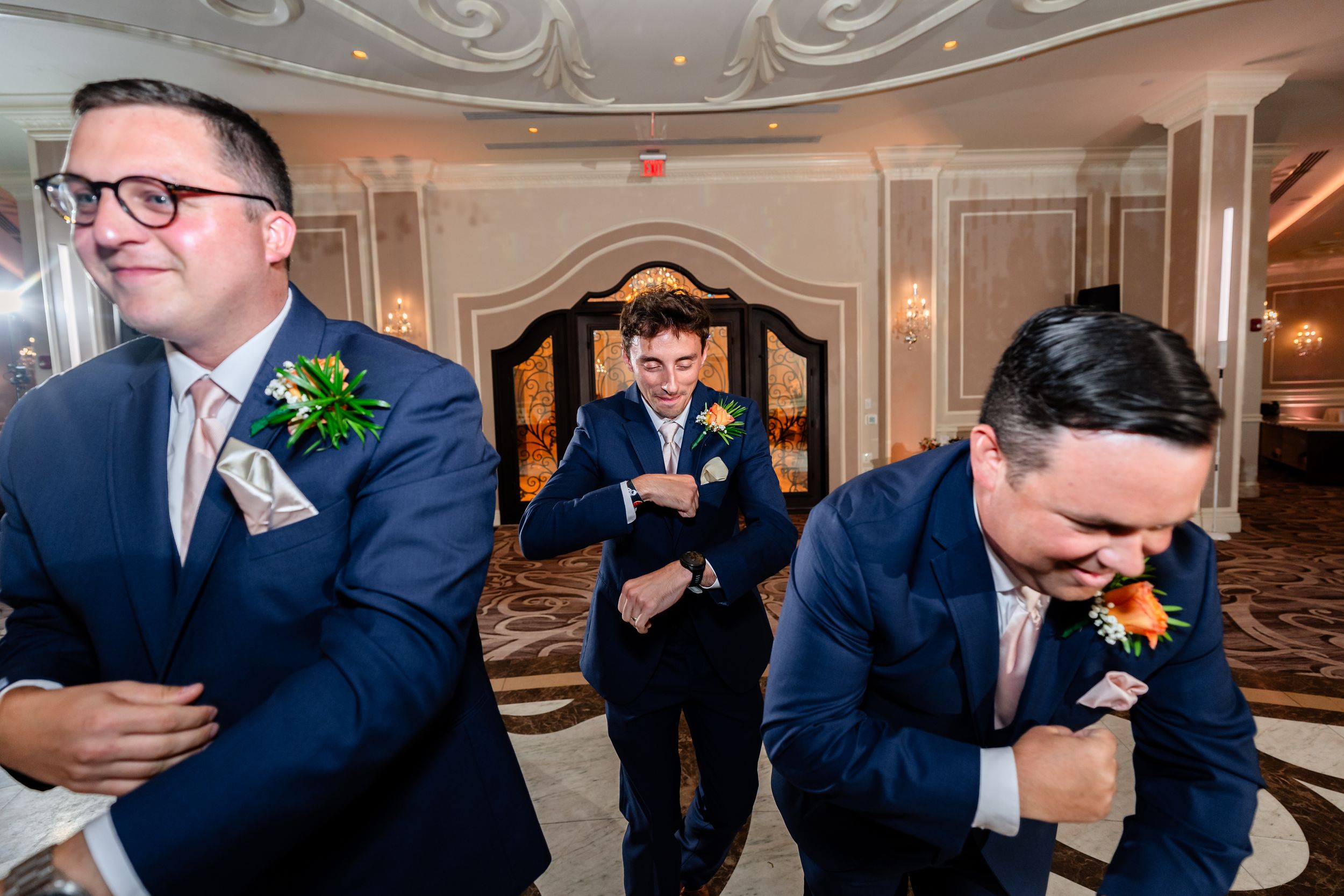 Three groomsmen in navy suits laugh and adjust their boutonnieres in an elegant ballroom.