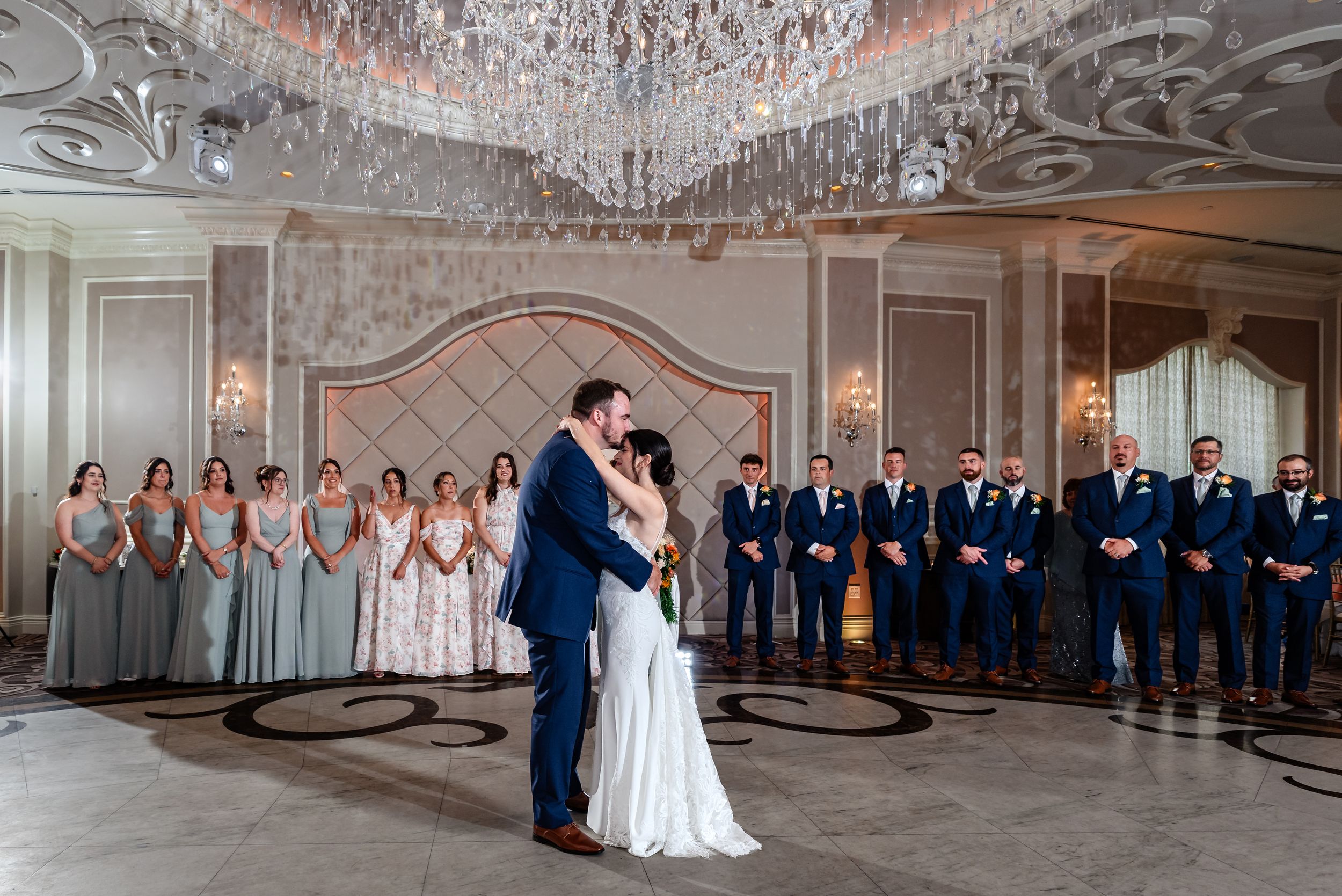 Bride and groom share first dance in elegant ballroom surrounded by wedding party under crystal chandelier.