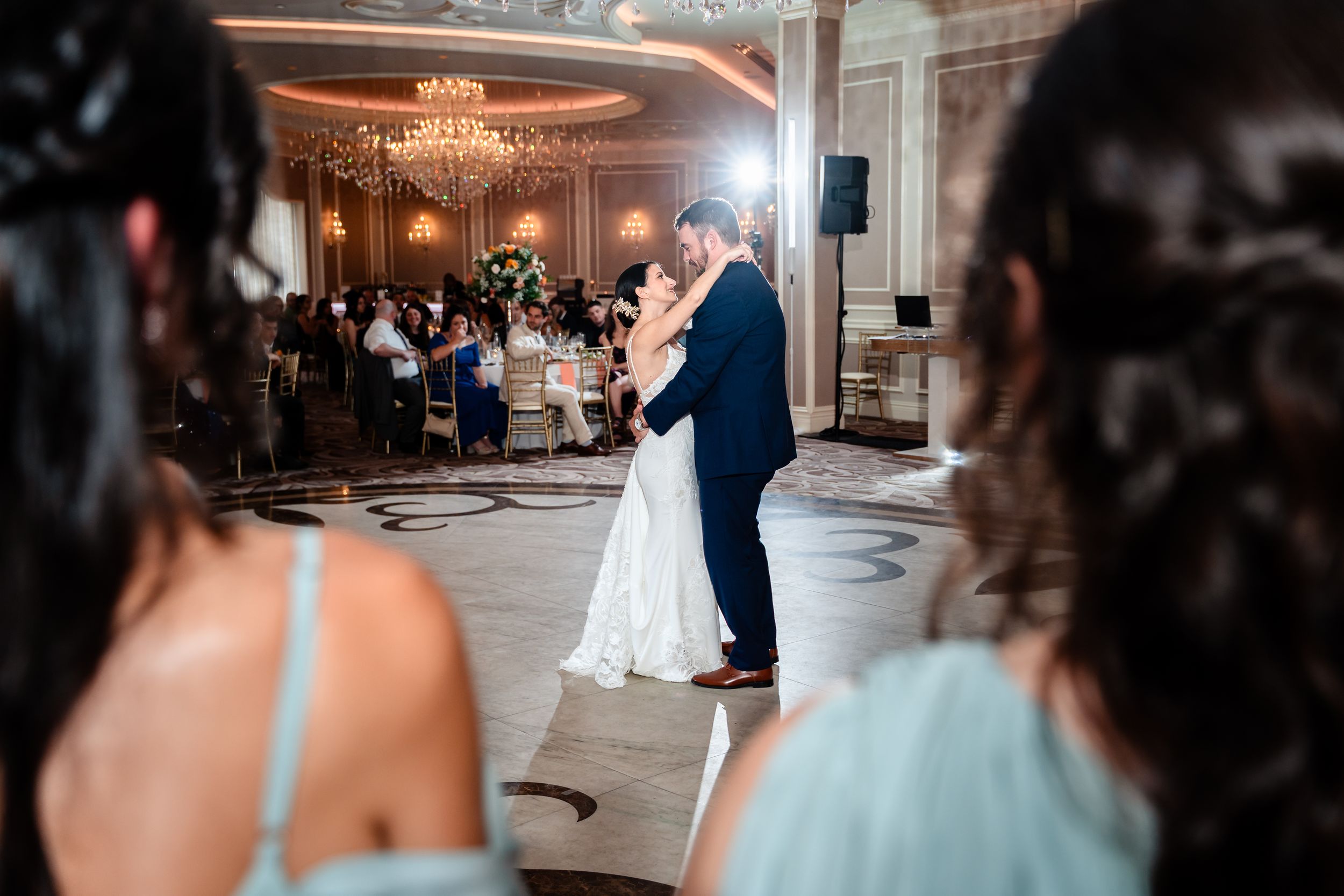 Bride and groom share their first dance in an elegant ballroom while wedding guests watch.