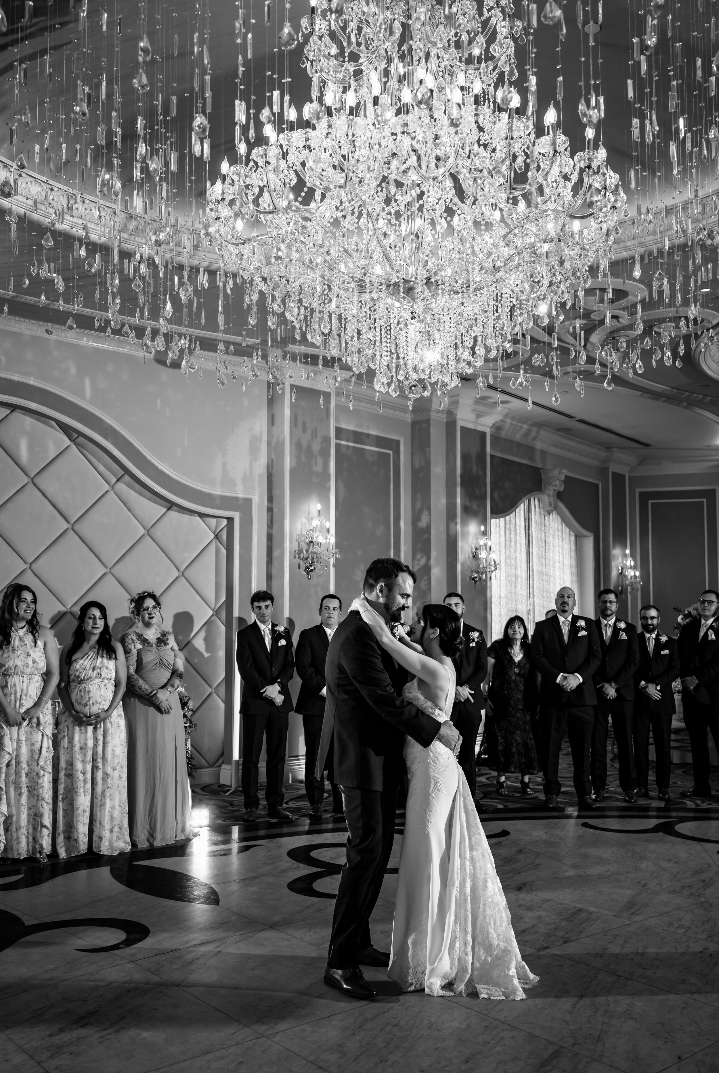 Bride and groom share first dance under grand crystal chandelier in elegant ballroom surrounded by wedding guests.