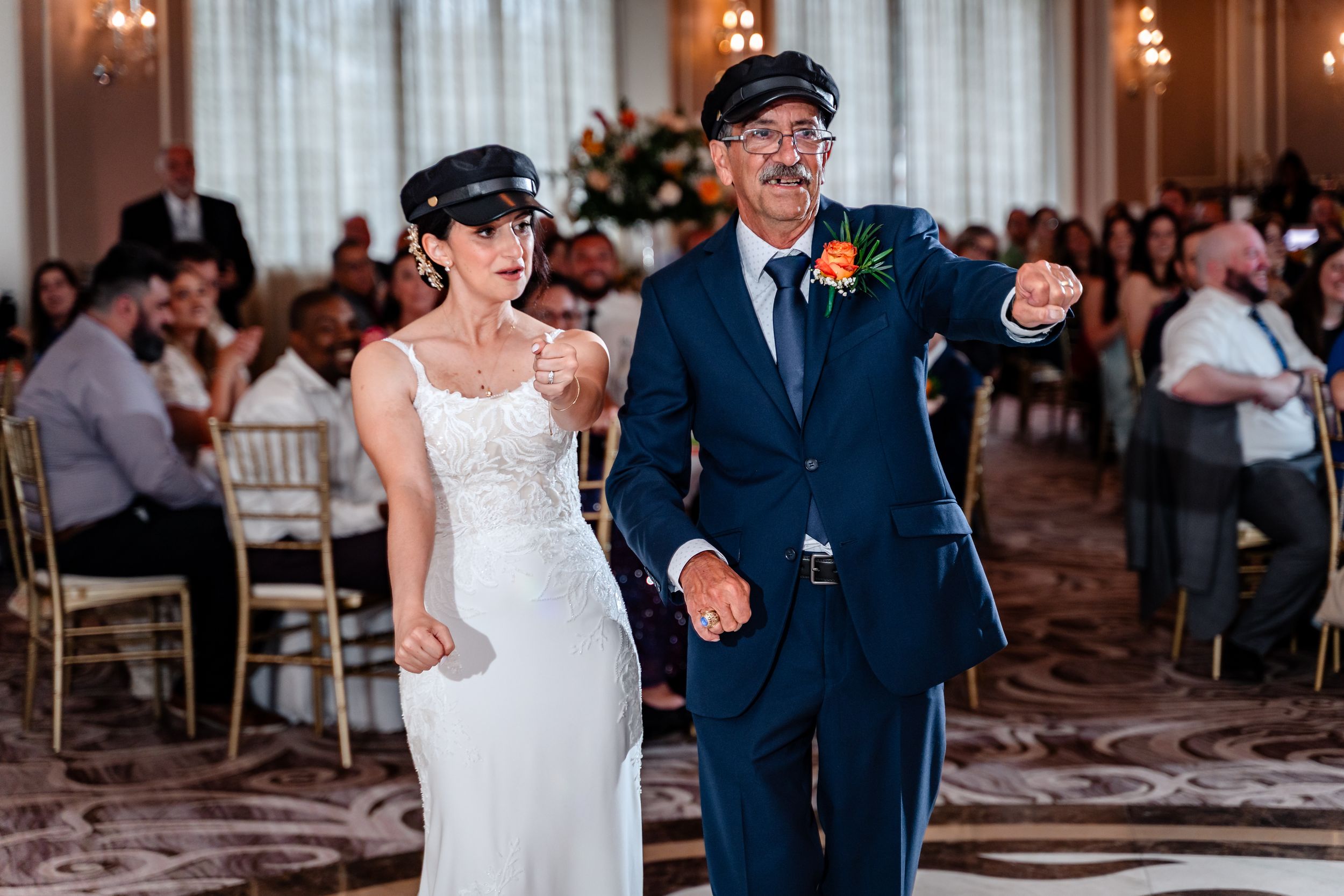 Bride in white gown and groom in navy suit wearing matching captain hats make a grand entrance at elegant wedding reception.