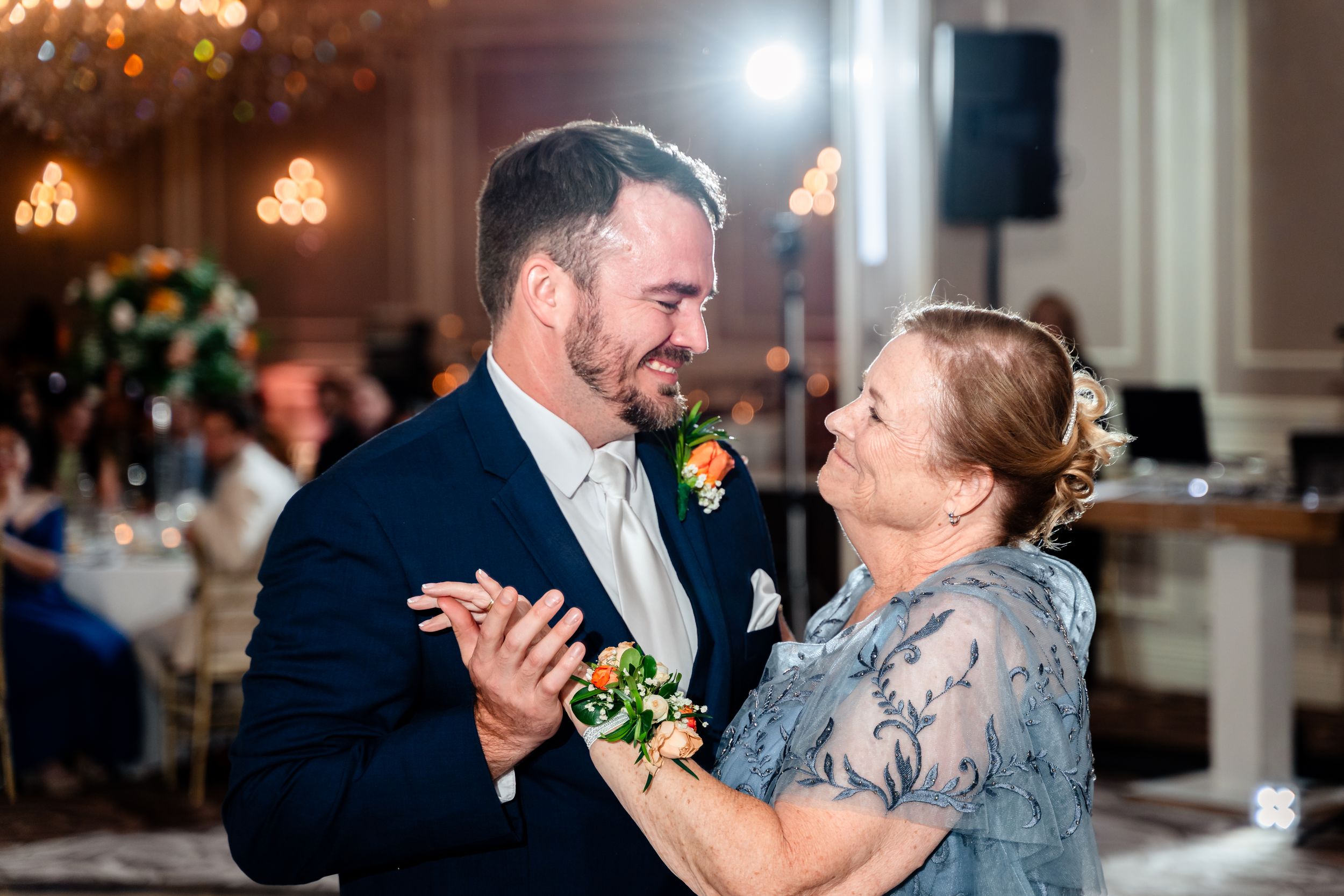 Groom in navy suit shares a tender mother-son dance at wedding reception.