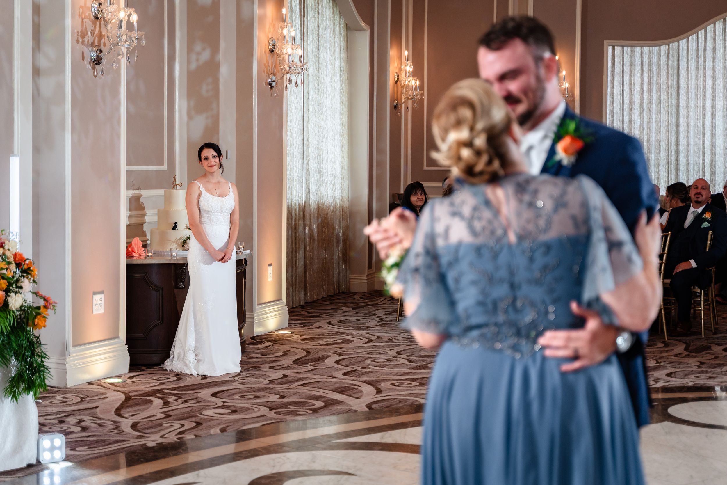 Bride watches groom share mother-son dance in elegant ballroom during wedding reception.