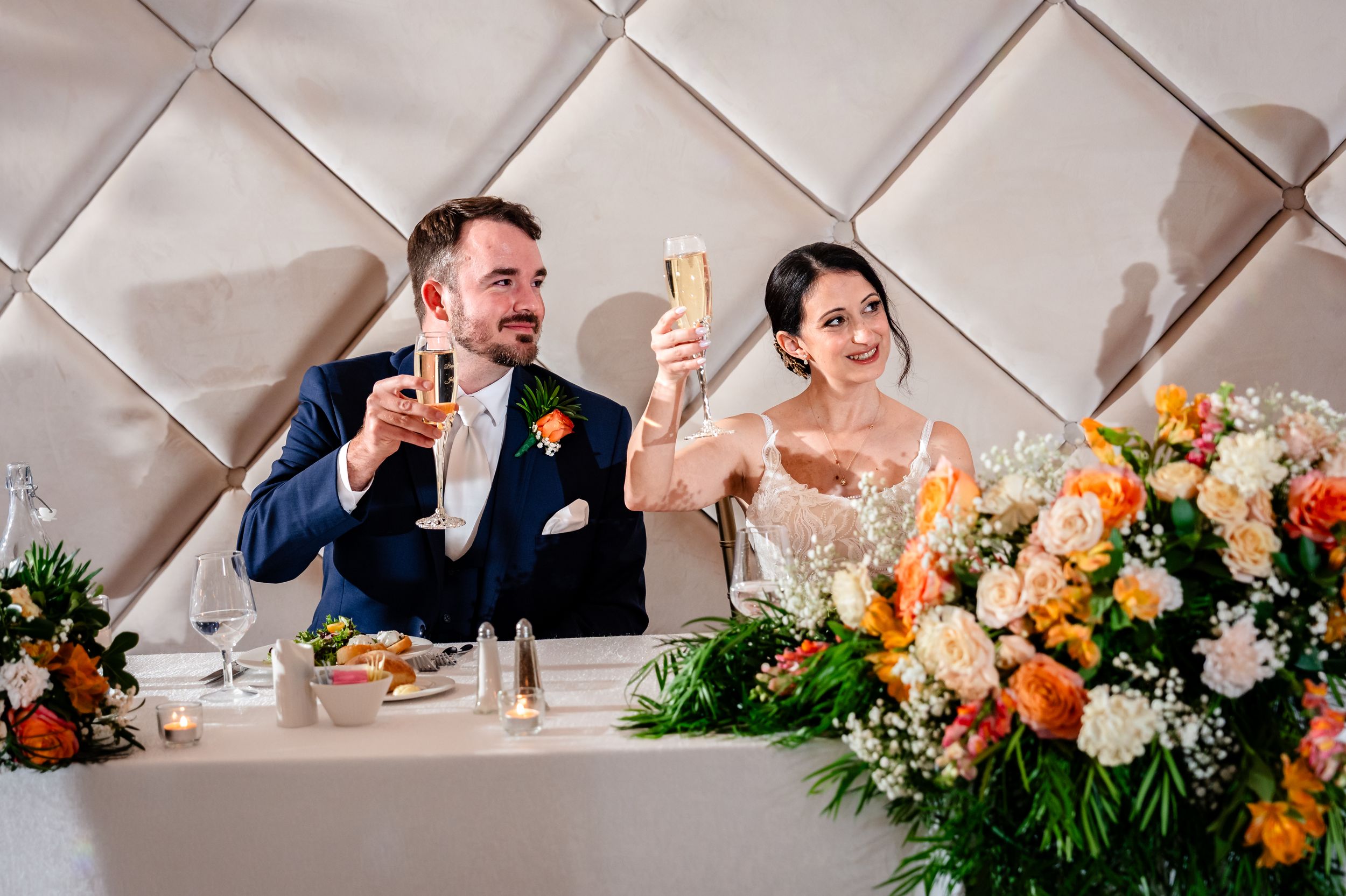 Bride and groom toasting with champagne at sweetheart table with floral arrangement.