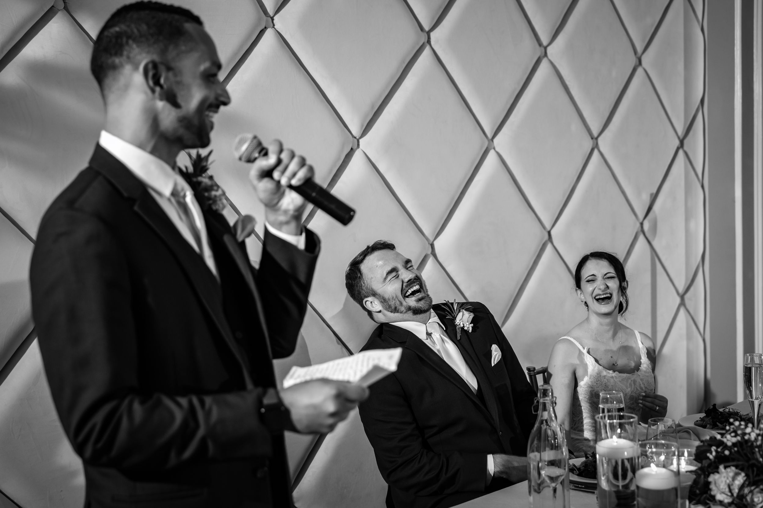 Black and white photo of a best man giving a humorous wedding speech while the groom and bride laugh joyfully.