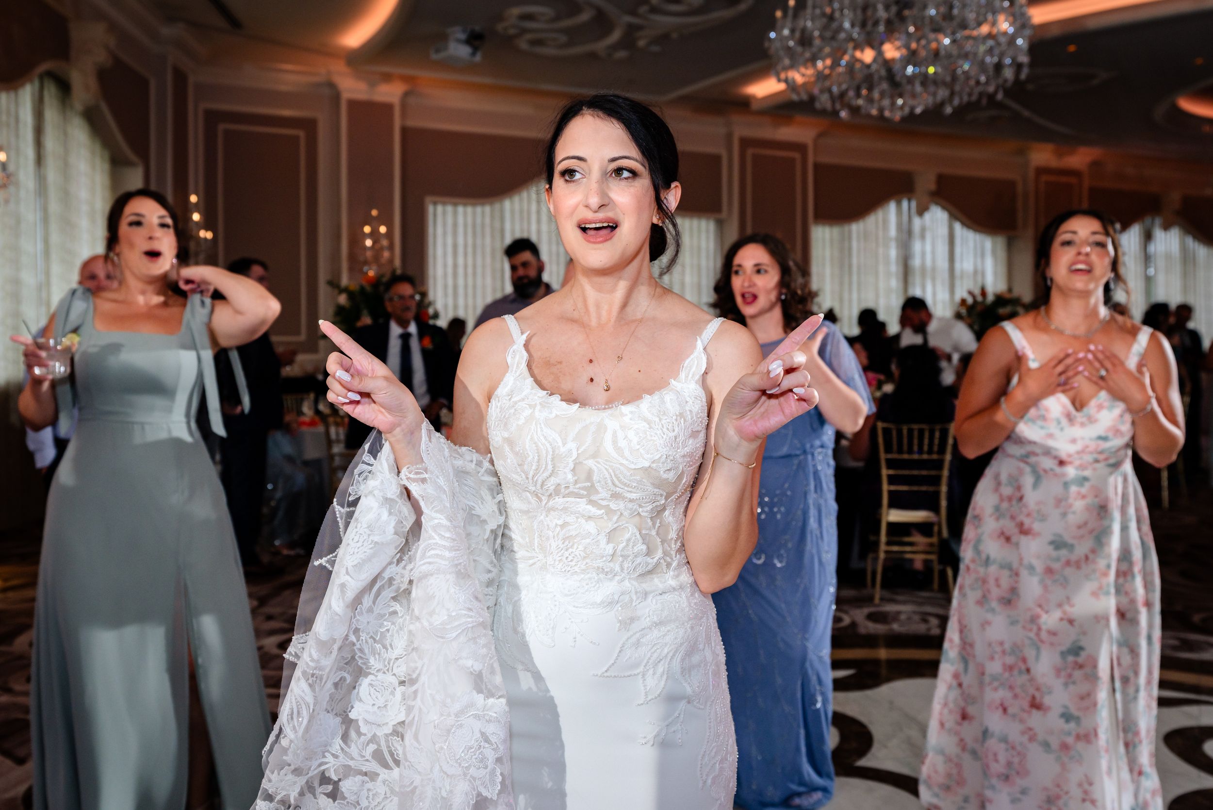 Bride in white lace wedding gown dancing joyfully with bridesmaids in elegant ballroom reception.