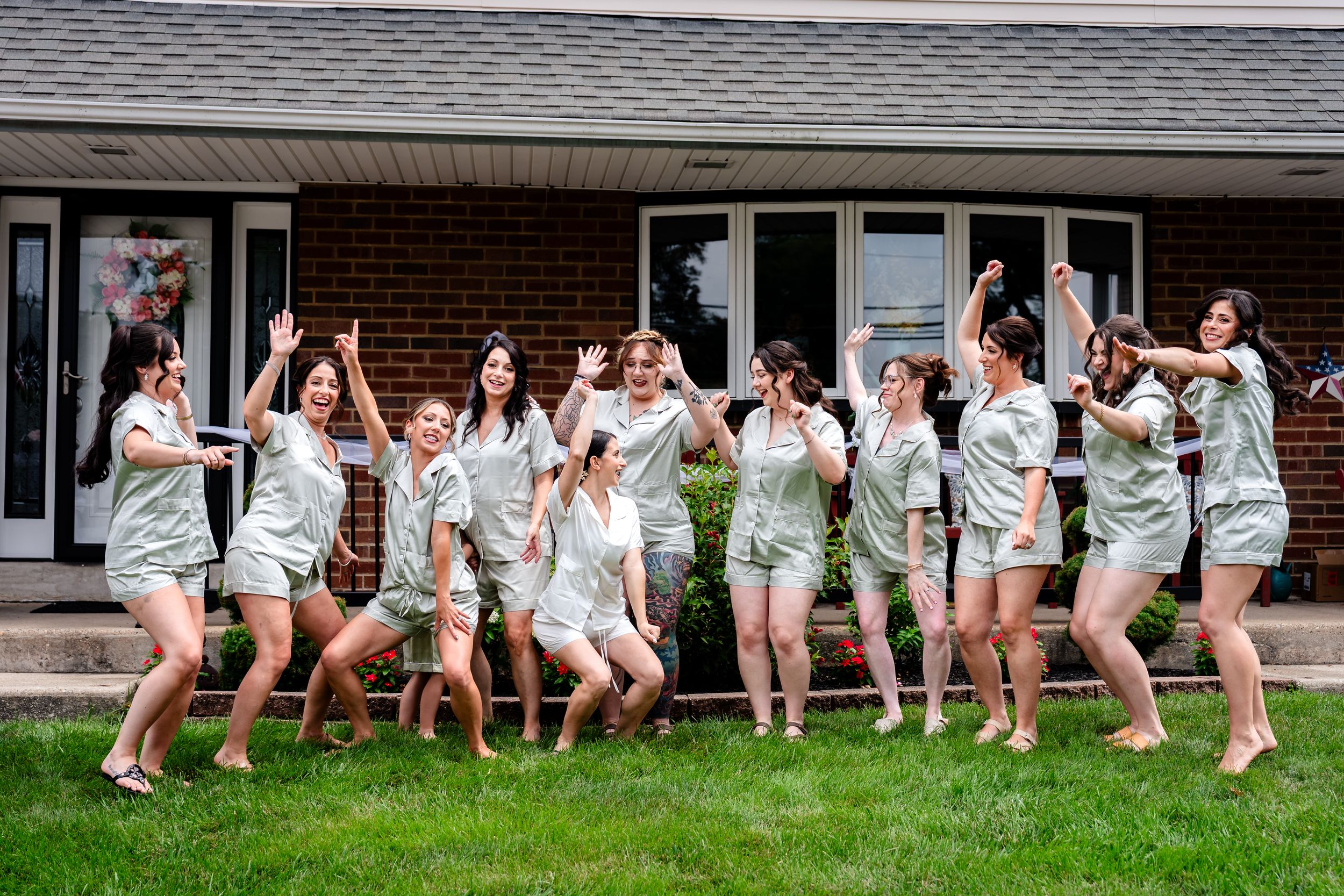 Bridesmaids in matching sage green pajamas posing and dancing on a lawn in front of a brick house.