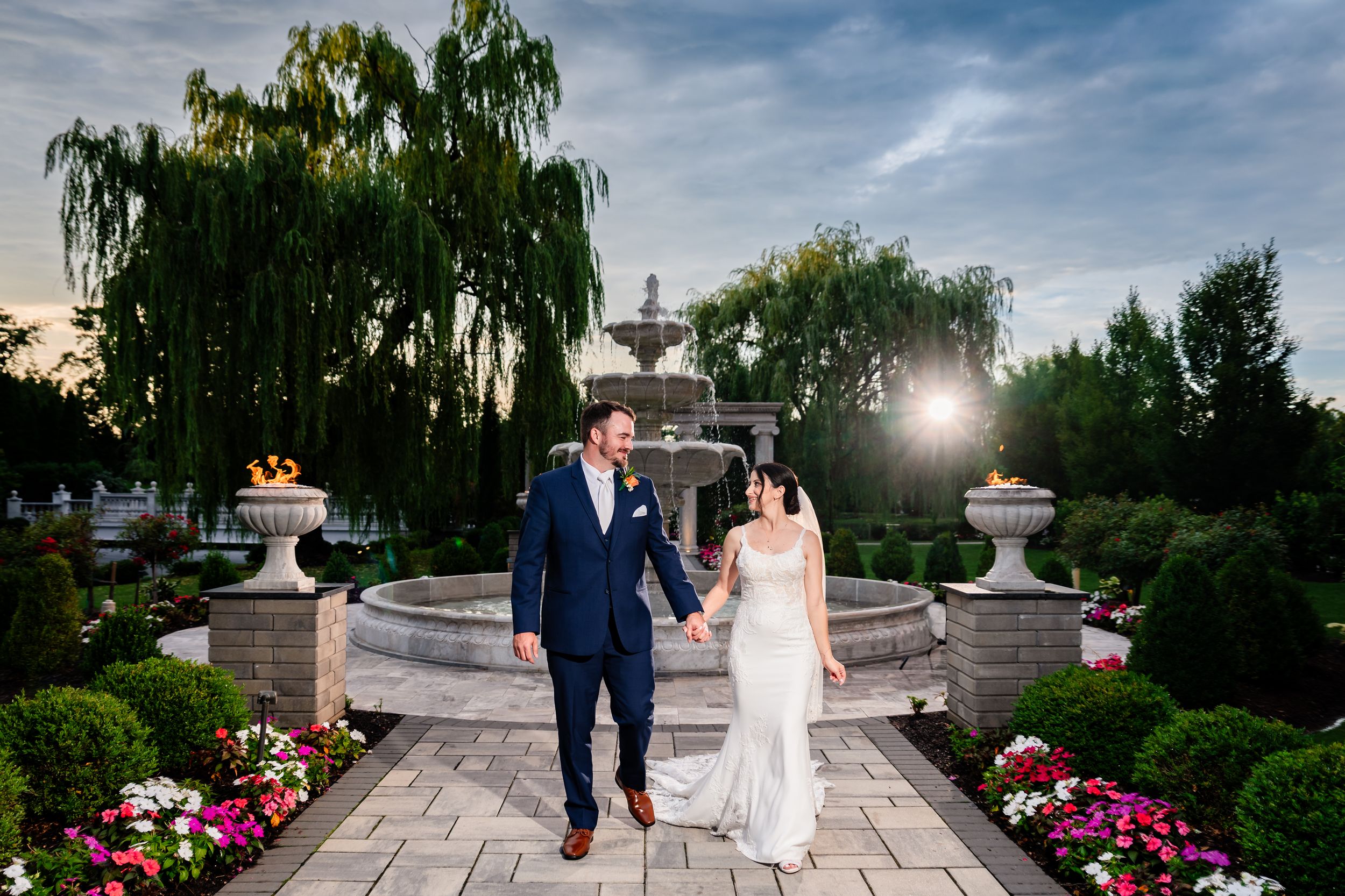 Bride and groom walk hand in hand through stunning garden with fountain at sunset.