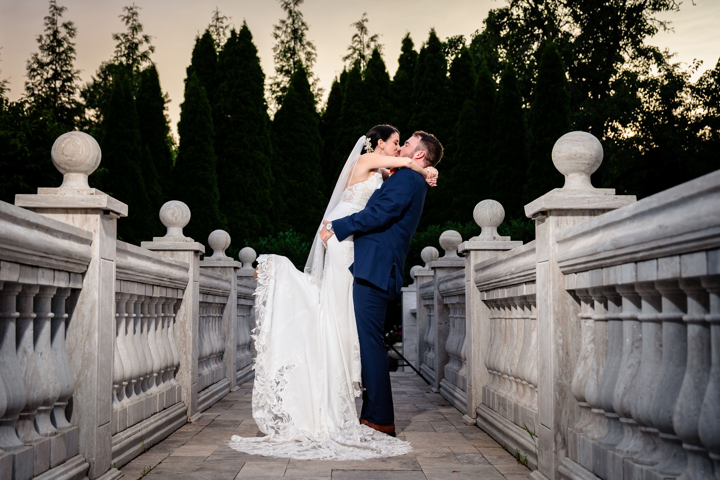 Bride and groom share romantic kiss on stone balcony at sunset surrounded by tall trees.