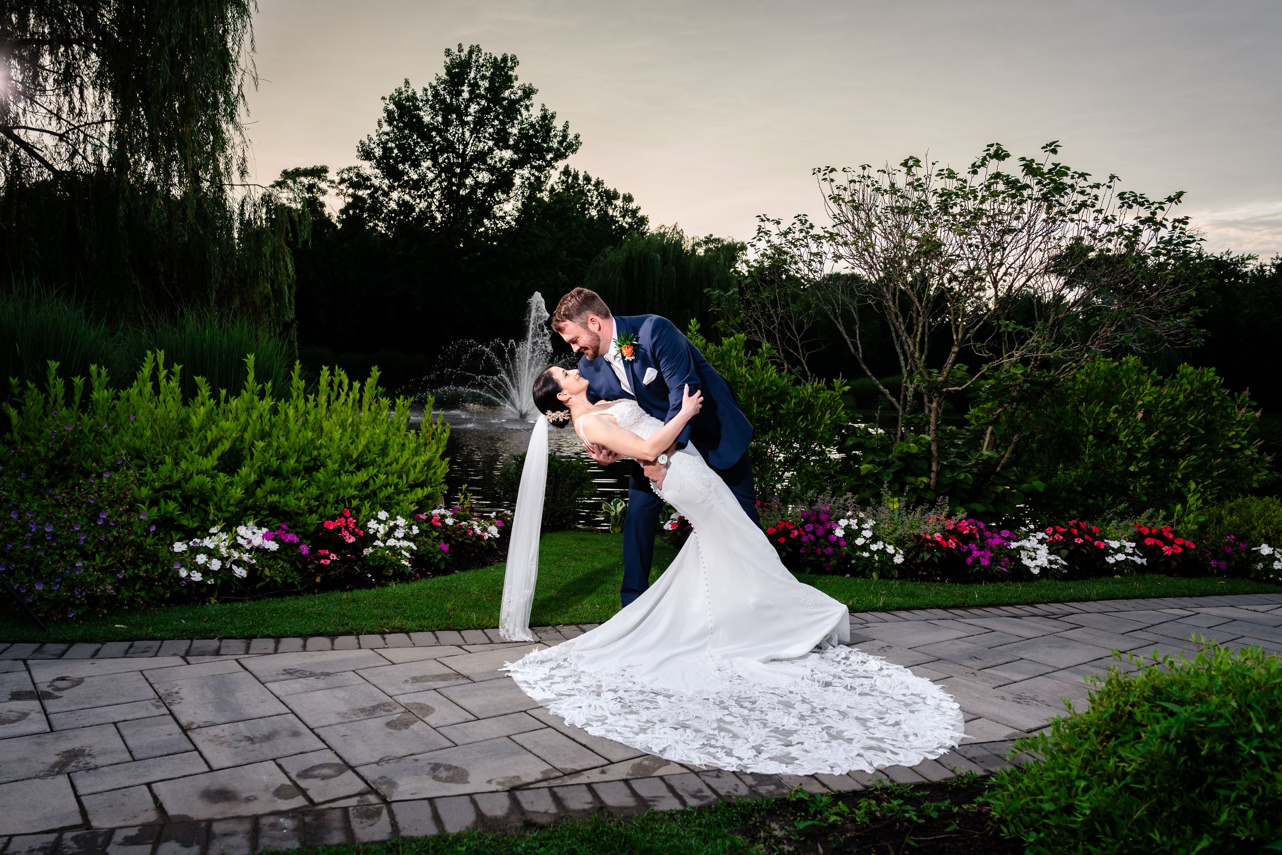 Groom dips bride in a romantic pose on a garden path surrounded by colorful flowers at sunset.