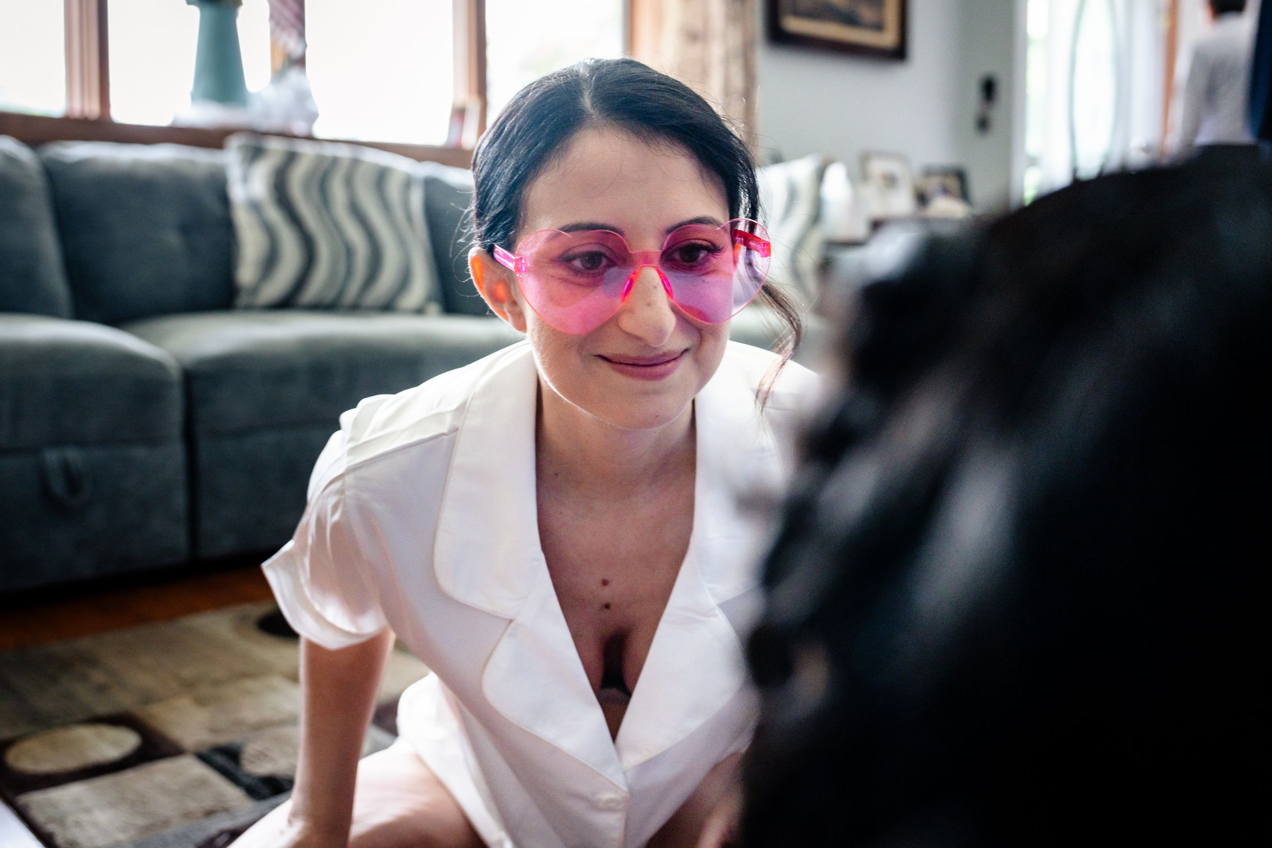 Young woman smiling wearing pink heart-shaped sunglasses sitting casually in a living room.
