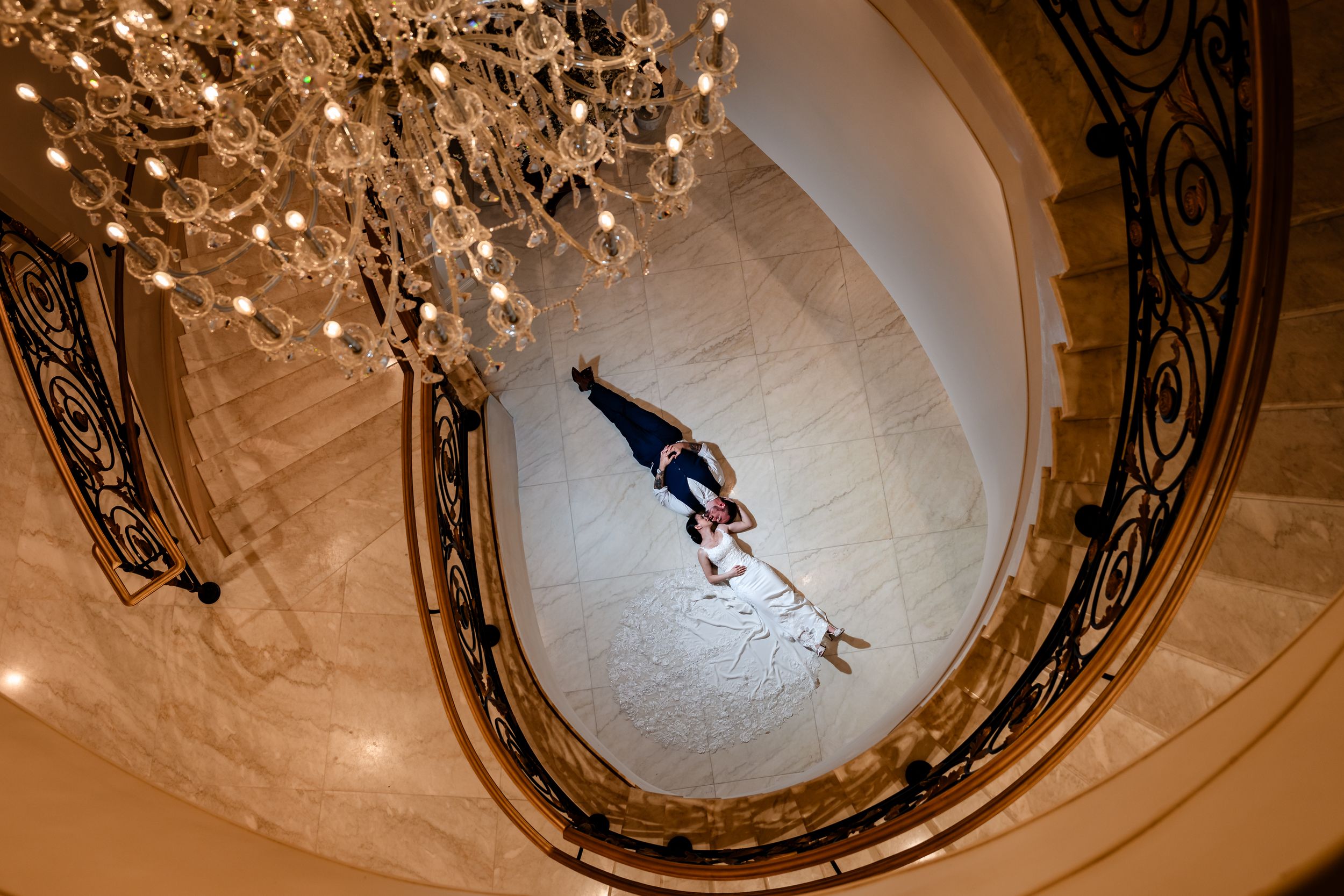 Aerial view of bride and groom posing on marble staircase beneath grand crystal chandelier.