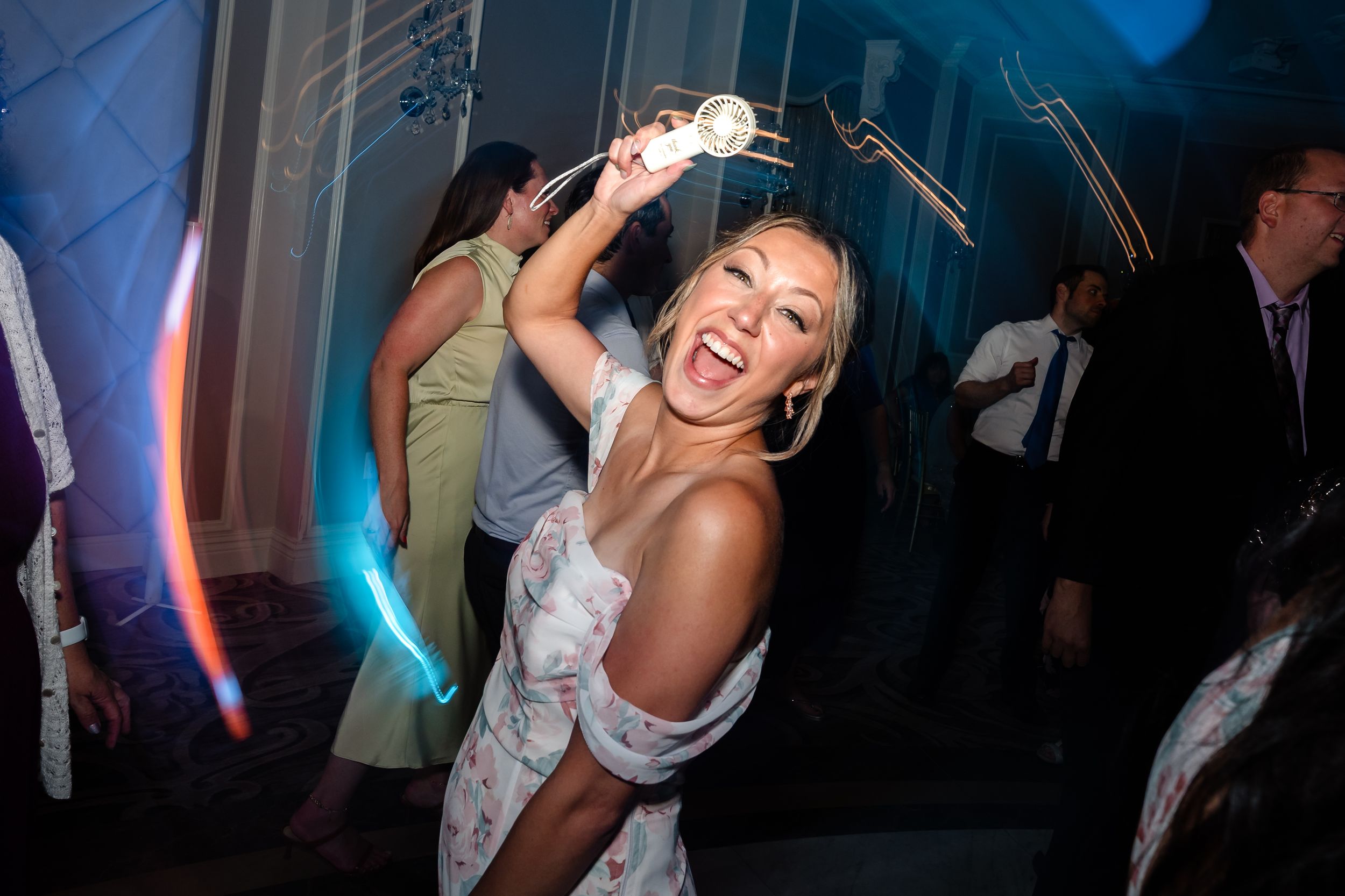 Joyful woman in floral dress dancing and laughing at a wedding reception party with colorful lights.
