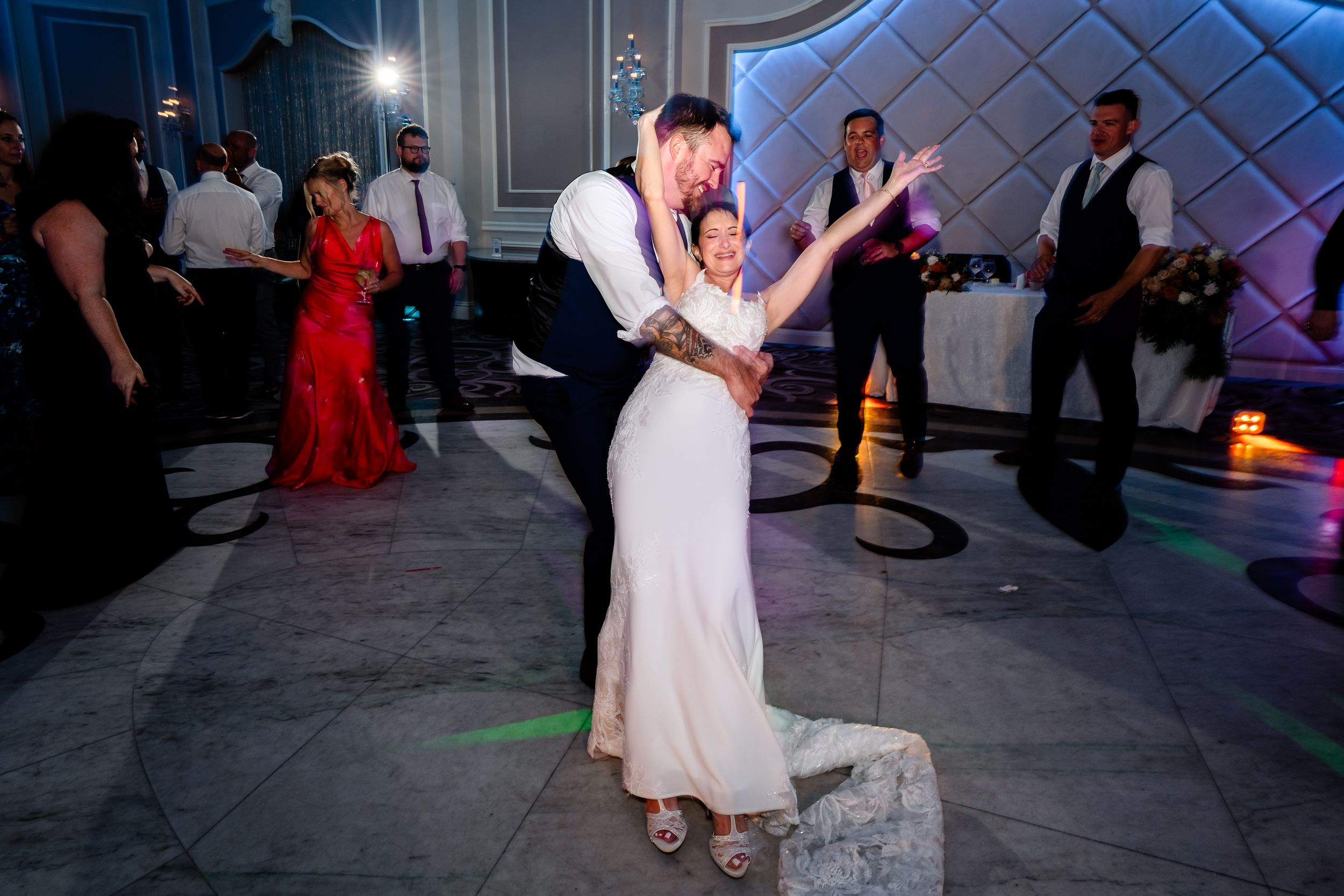 Bride and groom share joyful first dance surrounded by guests on lit wedding reception floor.
