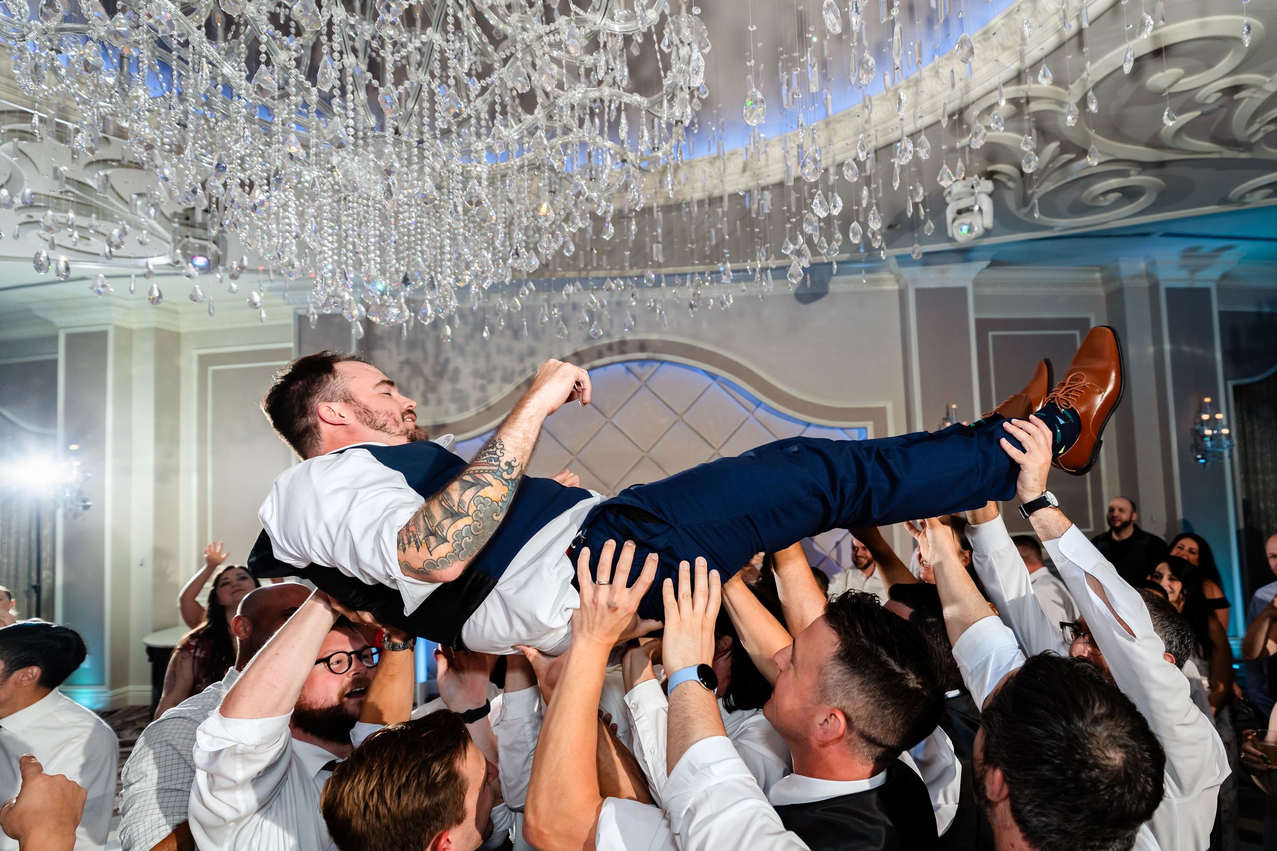 Groom being lifted by groomsmen at elegant wedding reception with crystal chandeliers.