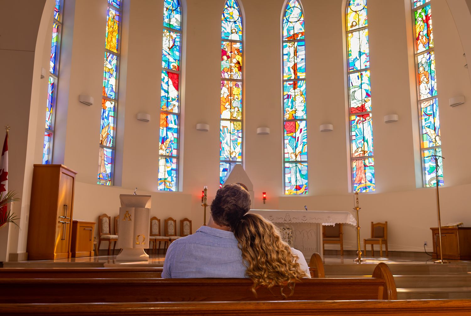 A couple sitting in a church pew, the woman resting her head on the man's shoulder while looking forward at the churches stained glass.