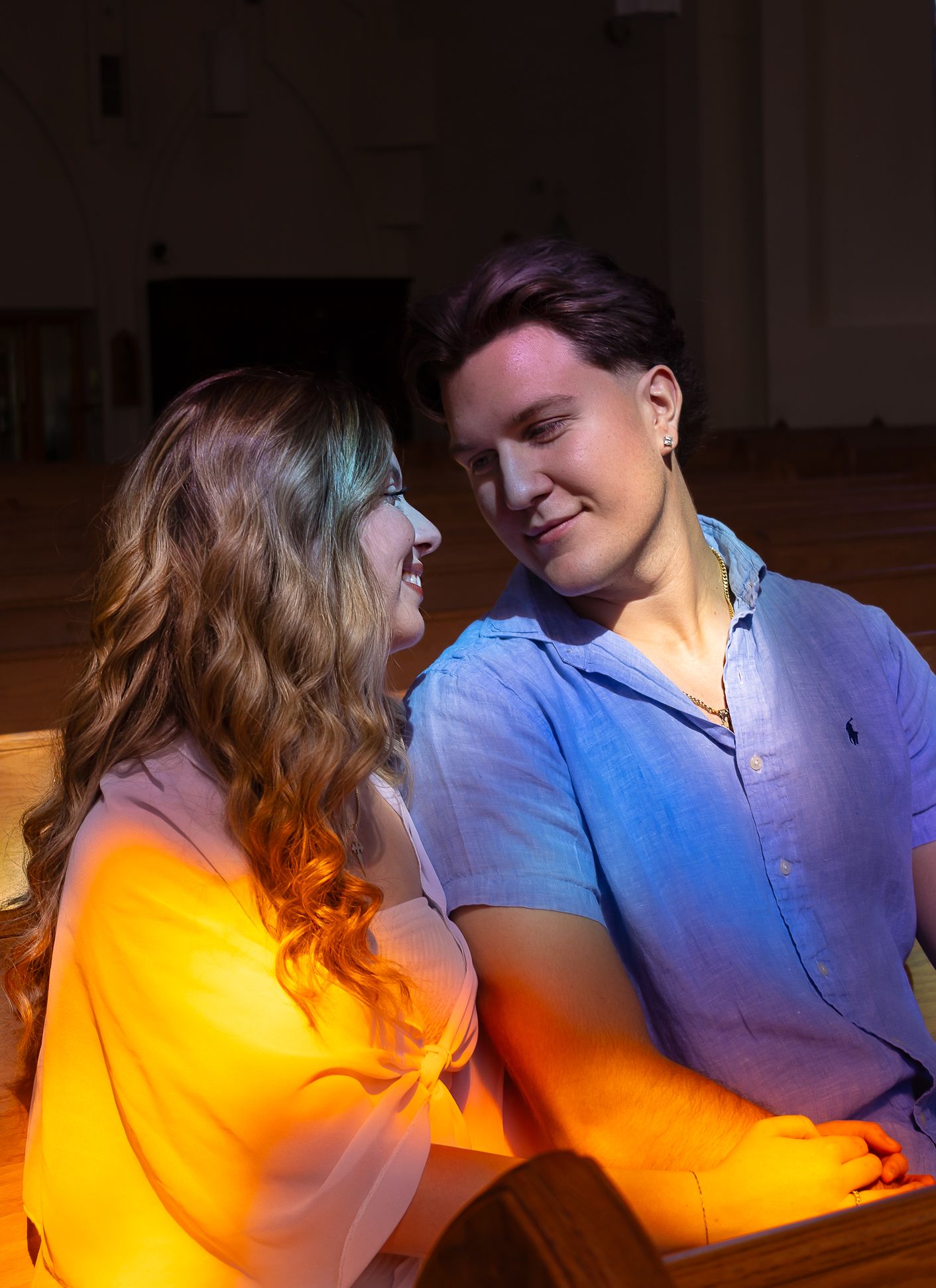 A woman and a man sitting in a Church pew, close up shot.  Staring into each other's eyes and smiling, with rainbow light hitting them