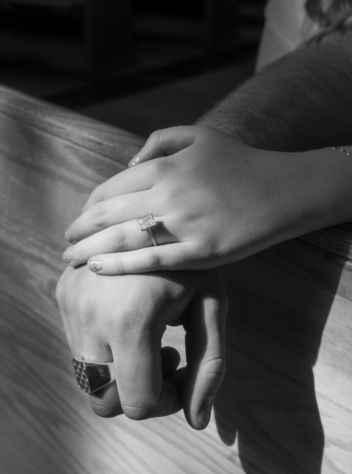 Black and white image of two hands, one on top of the other, resting on a church pew with an engagement ring in view