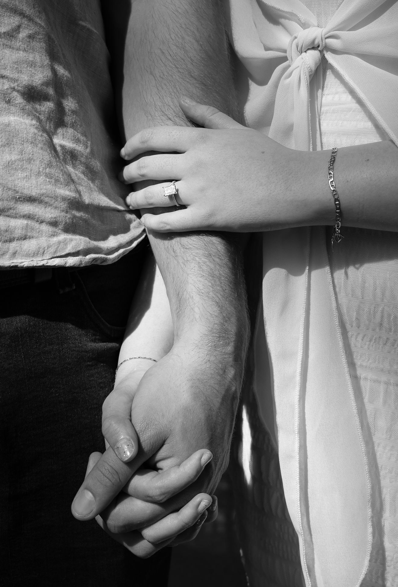 Black and white image of a couple holding hands, while the other hand rests on the forearm to show the engagement ring