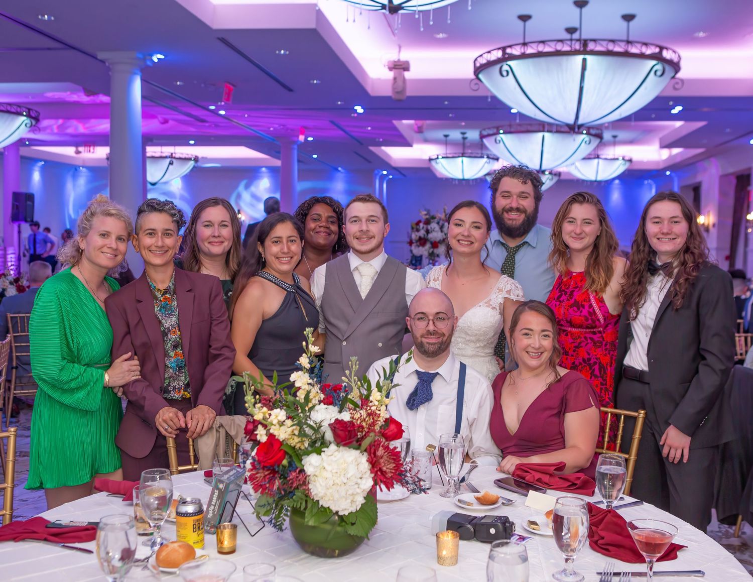 A large group of elegantly dressed guests poses together at a wedding reception in a beautifully lit ballroom.