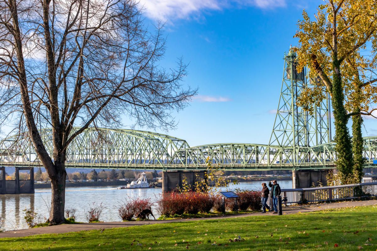 A green steel bridge spans a river beside a park with bare trees and autumn foliage on a sunny day.