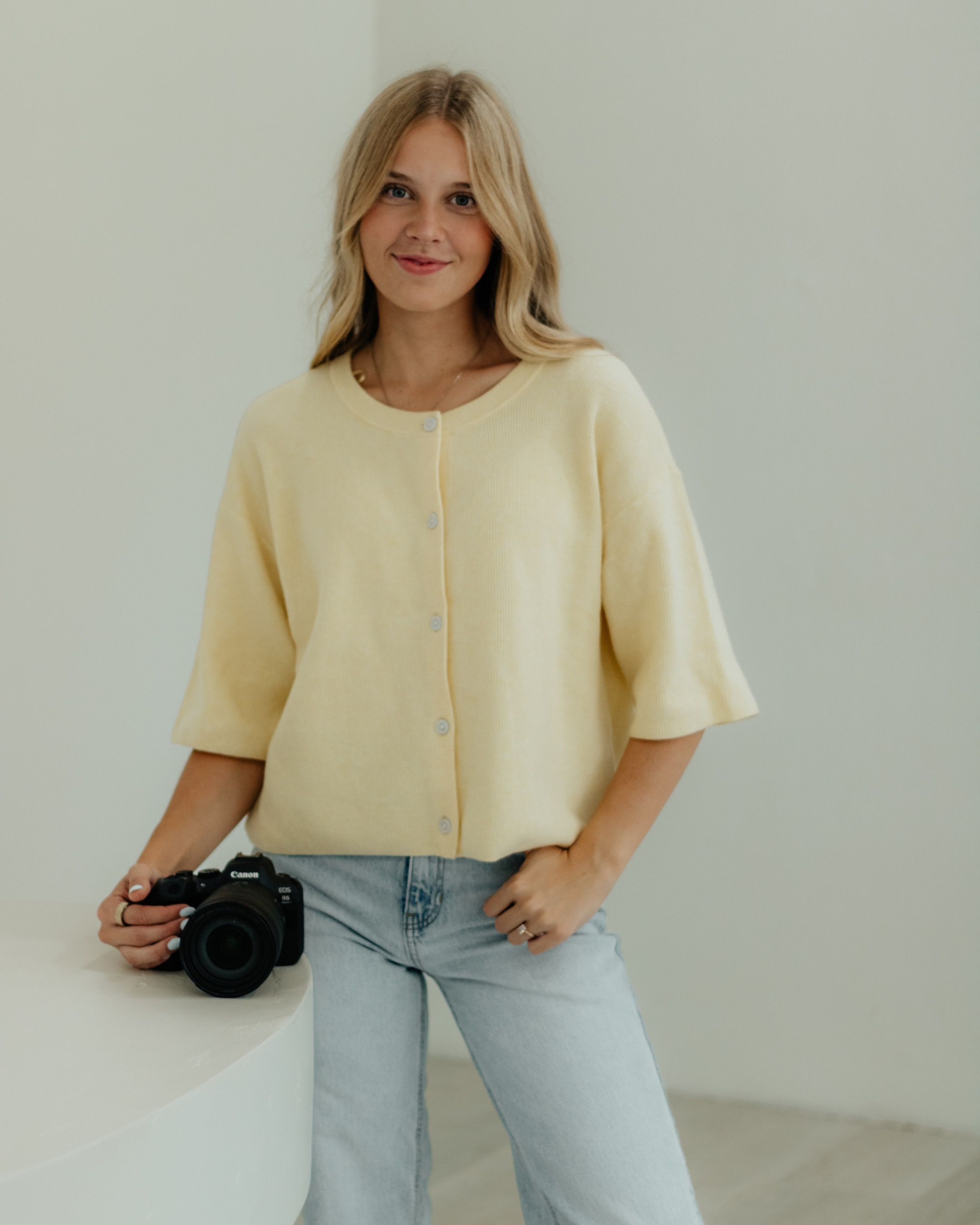 Woman in cream cardigan holding a Canon camera, wearing light blue jeans, posing against a white wall.