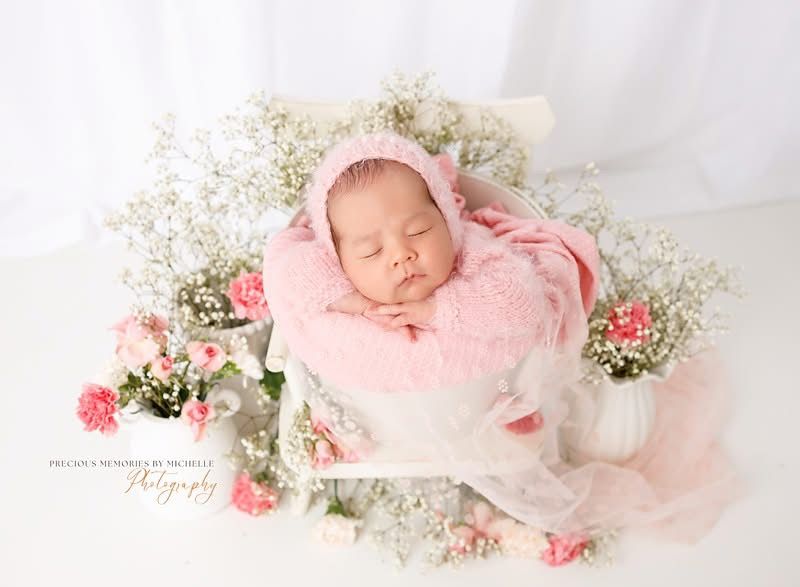 Newborn baby sleeping peacefully in pink bonnet on fluffy pink blanket surrounded by white and pink flowers.
