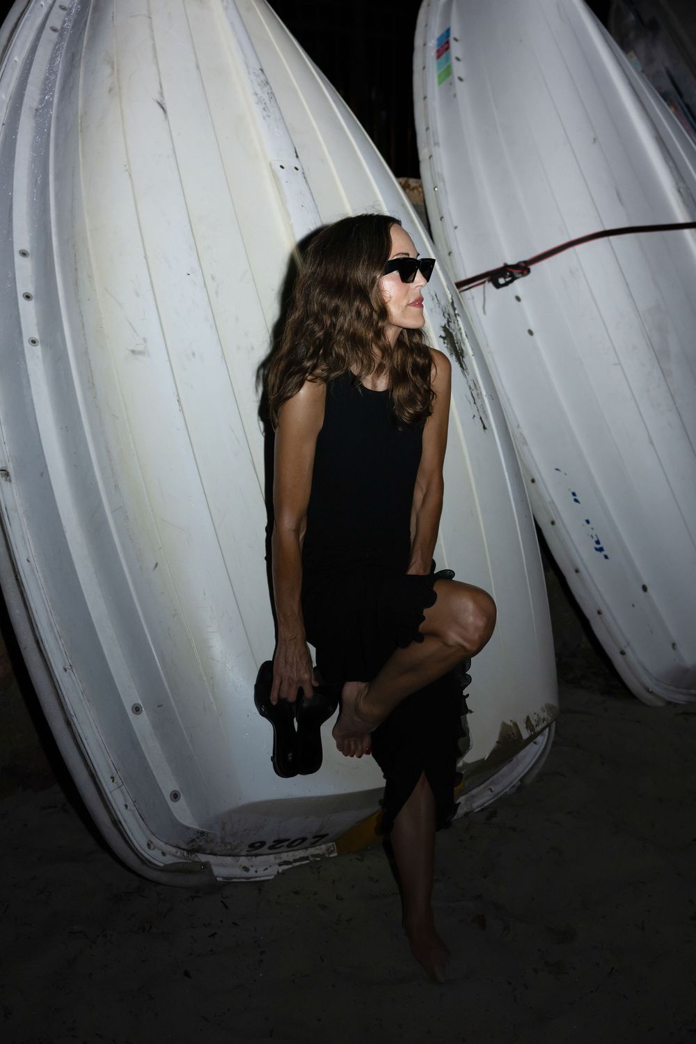 Woman in black dress and sunglasses posing barefoot against white boats stored outdoors at night.
