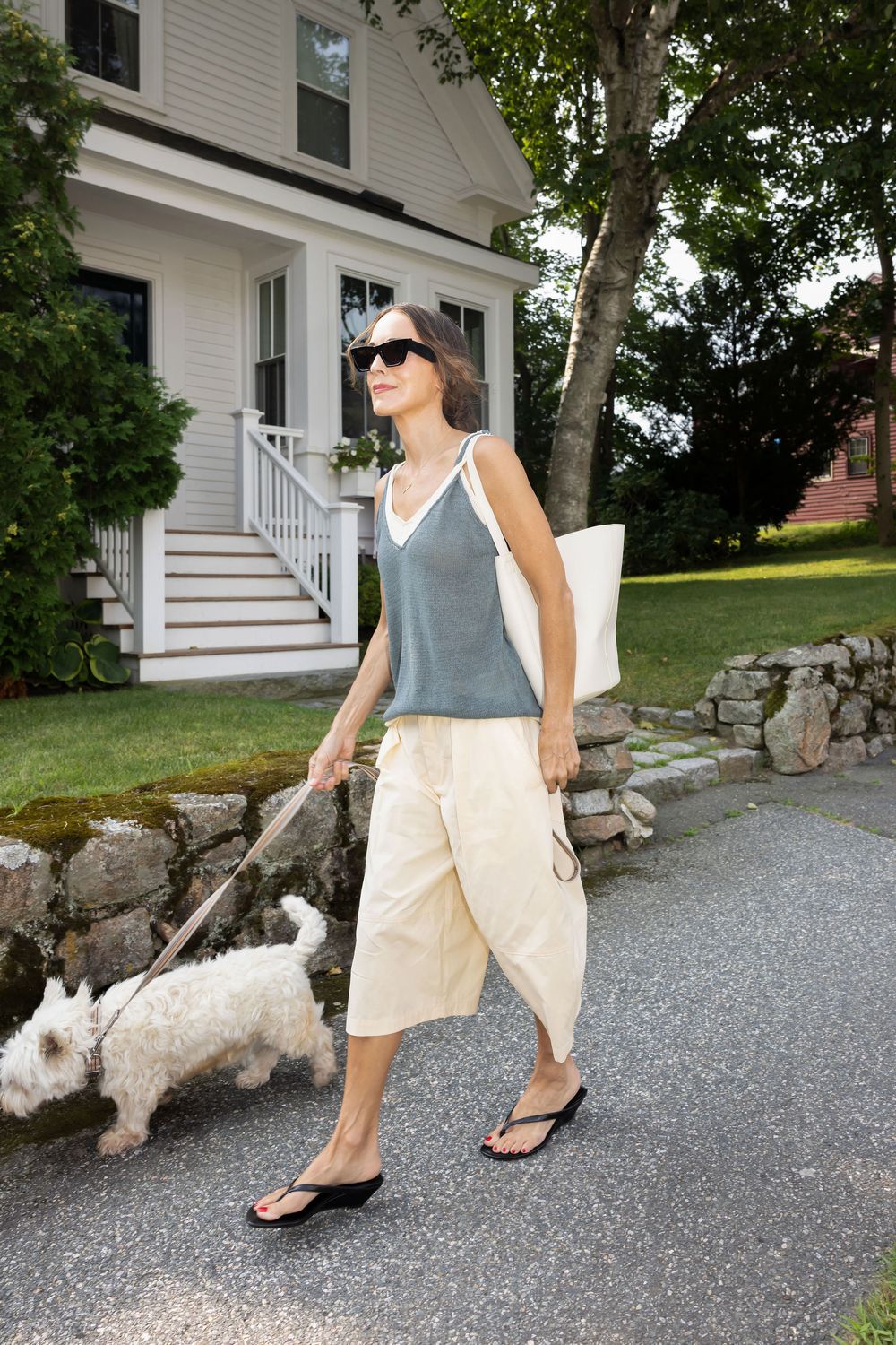 Woman in gray tank top and cream capri pants walks small white dog on leash past a white colonial house.