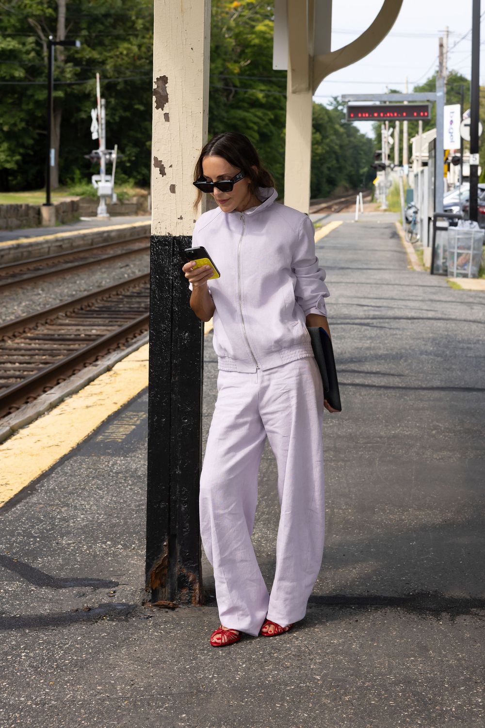 Woman in white linen suit with red flats checks phone at train station platform.