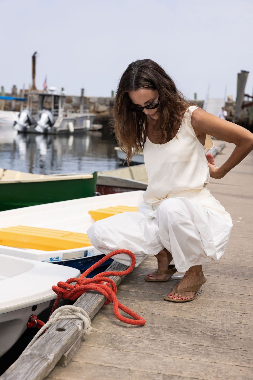 Woman in white outfit crouching on marina dock beside colorful boats with red rope.