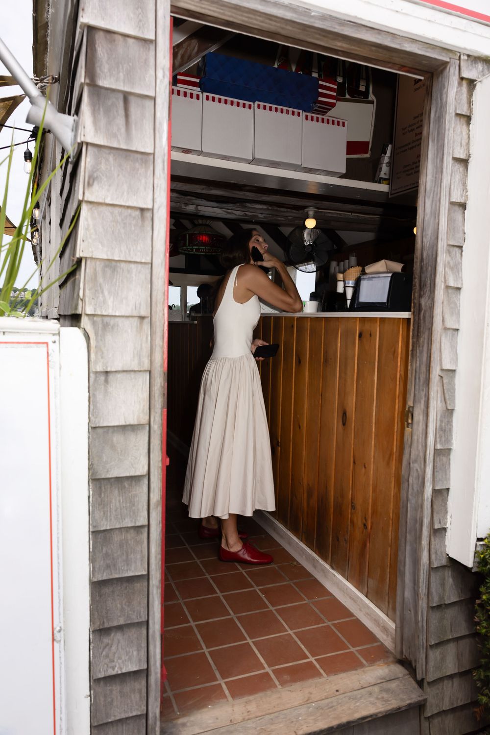 Woman in white midi dress and red flats standing in doorway of rustic wooden building.