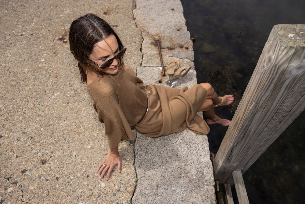Woman in tan outfit sitting on stone dock edge by water, viewed from above, wearing sunglasses.