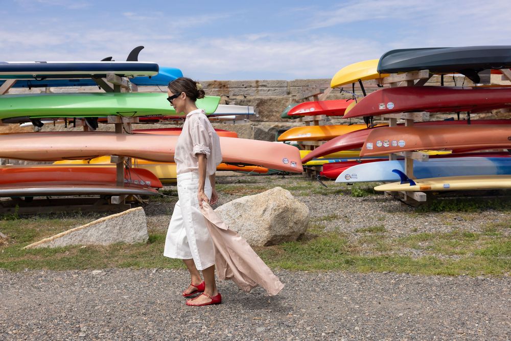 Woman in white dress walks past colorful kayaks stacked on outdoor racks near a rocky shoreline.