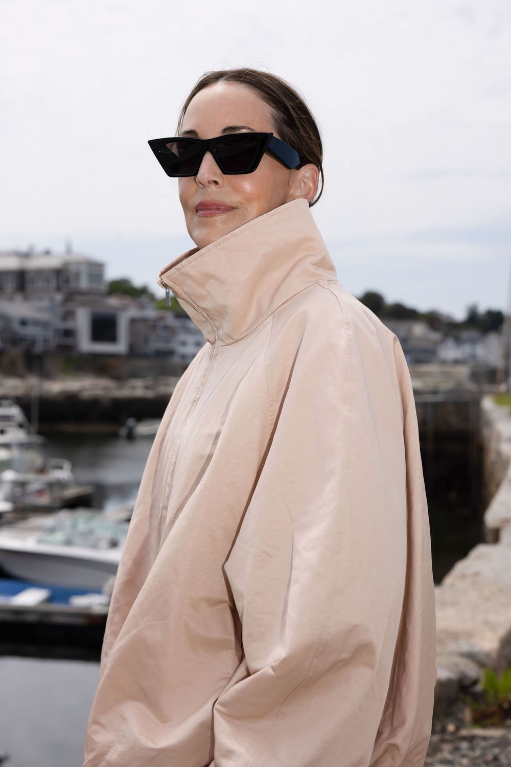 Woman in beige oversized jacket and black cat-eye sunglasses posing near a coastal harbor.