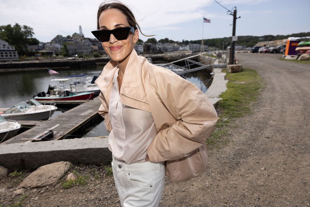 Woman in beige jacket and white jeans wearing cat-eye sunglasses poses near a marina on a sunny day.